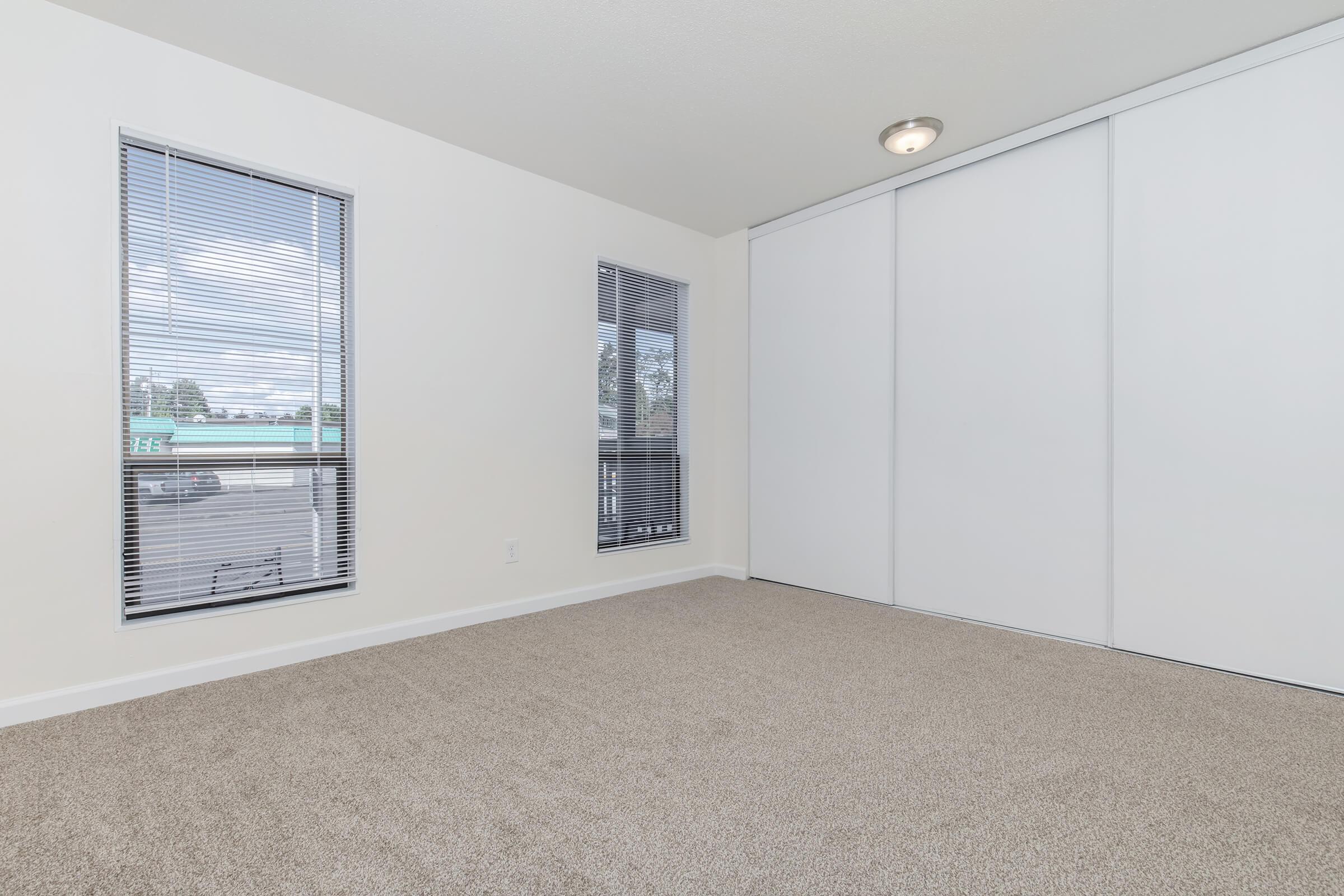 A spacious, brightly lit room with beige carpet flooring. It features two windows with blinds allowing natural light to enter, and one wall has sliding white closet doors. The walls are painted a light color, enhancing the open feel of the space. A ceiling light fixture is visible.