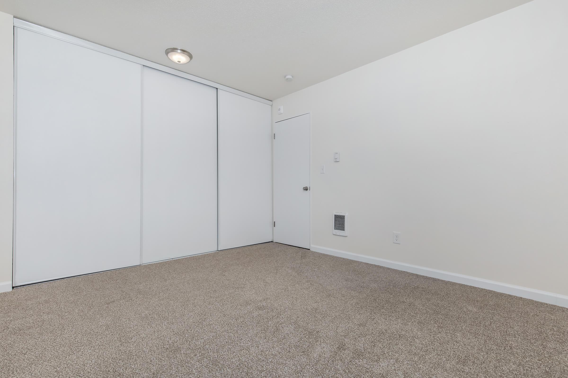 A spacious, empty room with light beige carpet, featuring a closed white wardrobe on one wall, a plain door on another, and a small vent near the floor. The walls are painted white and the ceiling has a simple light fixture. The room has a clean, modern look, ready for furnishings.