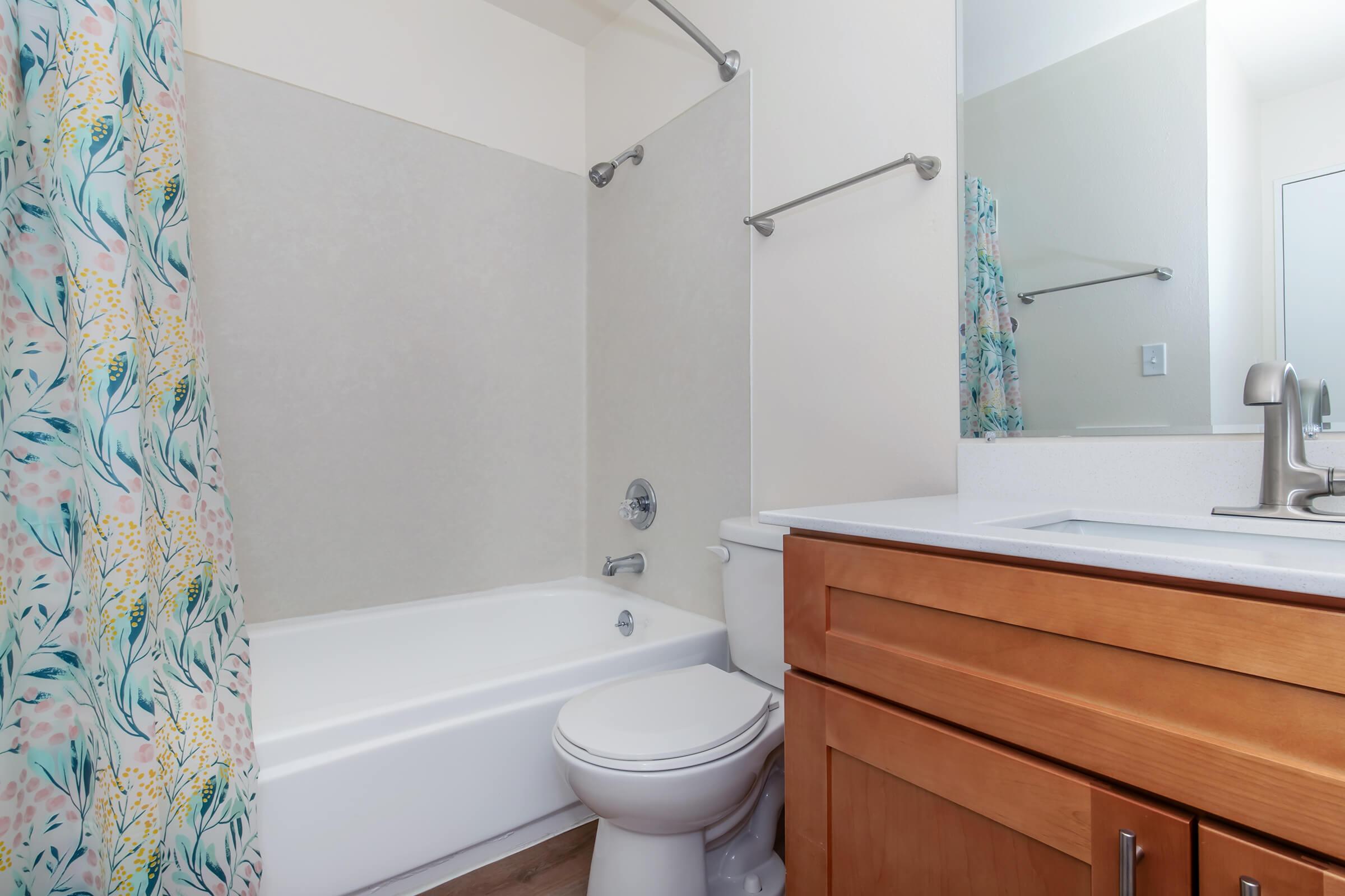 A clean bathroom featuring a white bathtub with a shower, a light-colored wall, and a wooden vanity with a sink. A colorful floral shower curtain adds a decorative touch. The bathroom also includes a modern mirror and a chrome faucet.