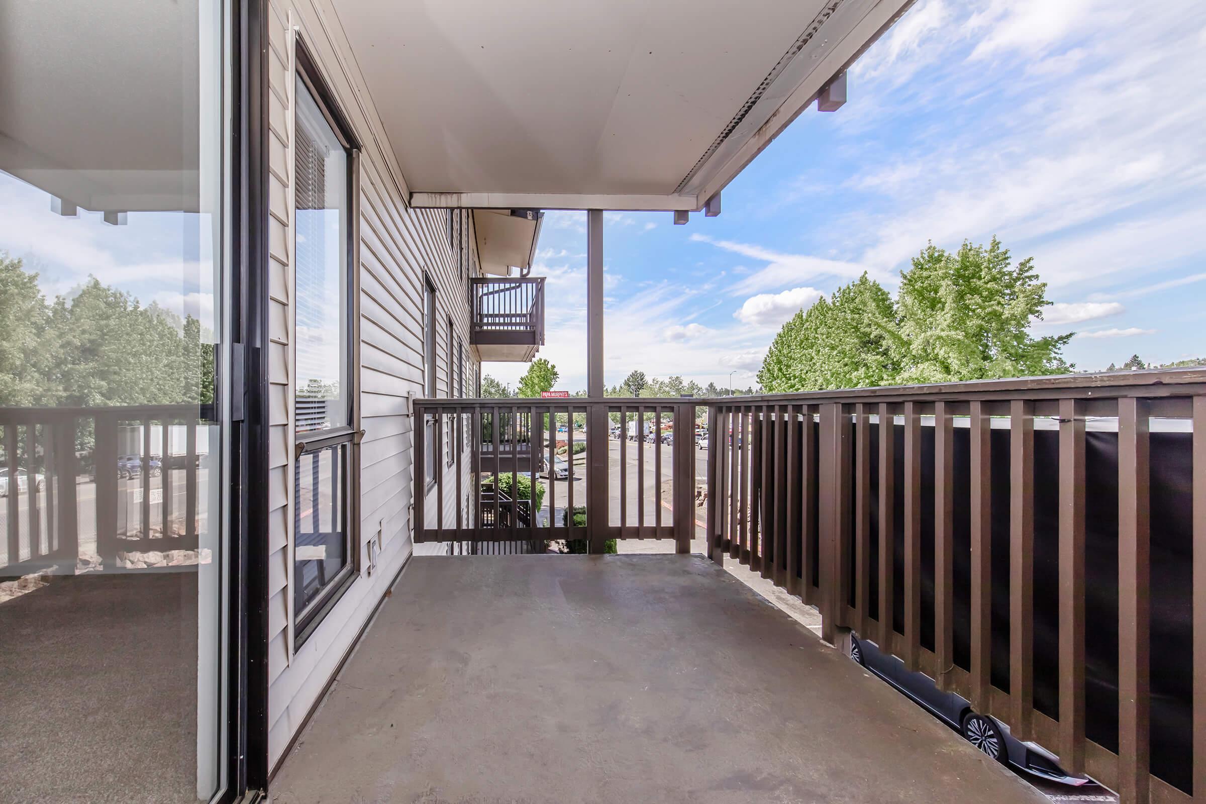 A balcony view featuring a concrete floor, wooden railing, and a glimpse of trees and blue sky in the background. The area is well-lit, showcasing a clear and spacious outdoor space.