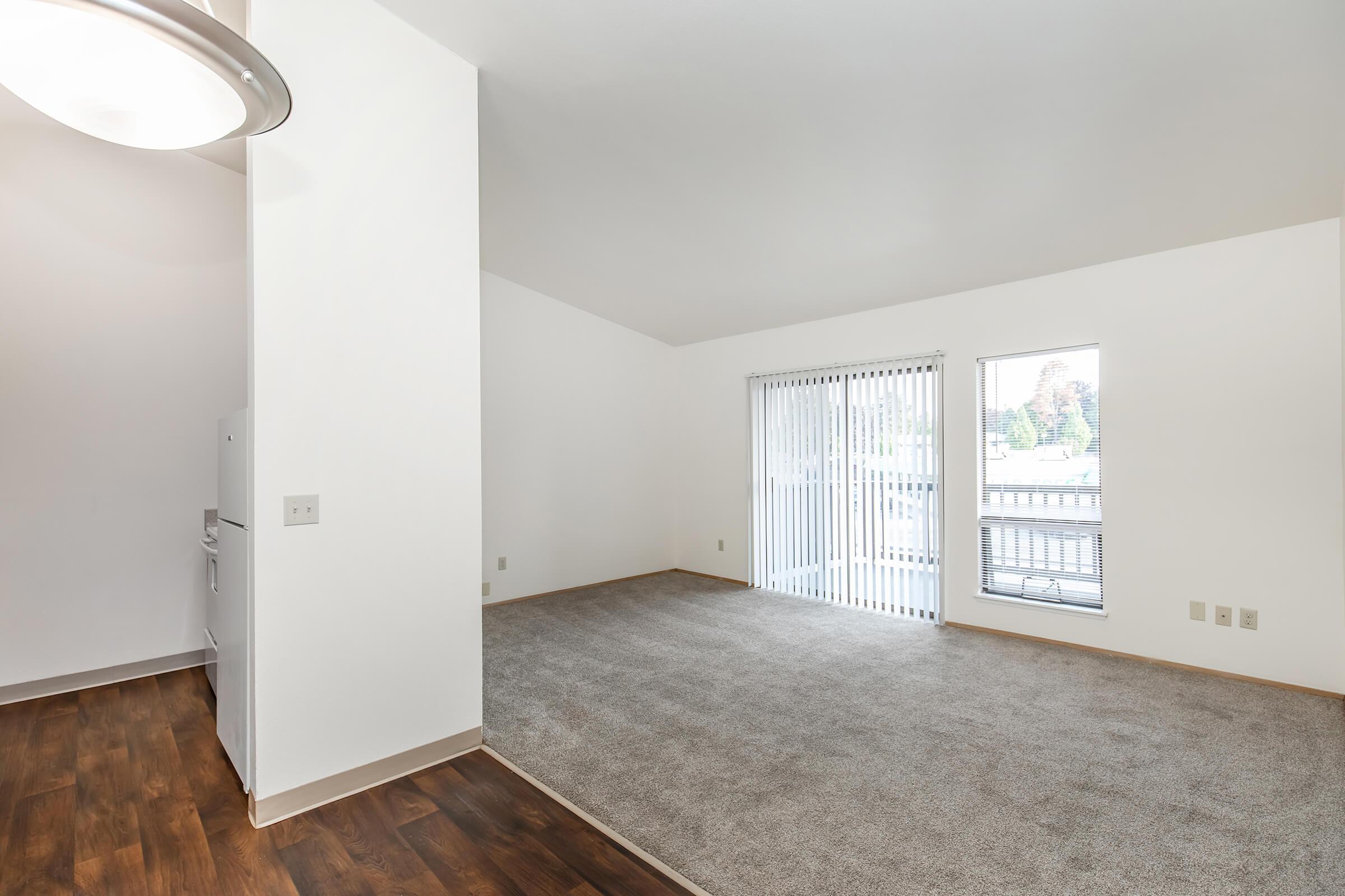 Spacious living area with high ceilings, featuring light-colored walls and a large window with vertical blinds. A doorway leads to a kitchen area on the left, and the floor is covered in plush, light brown carpet. The room is well-lit with natural light coming from the window.