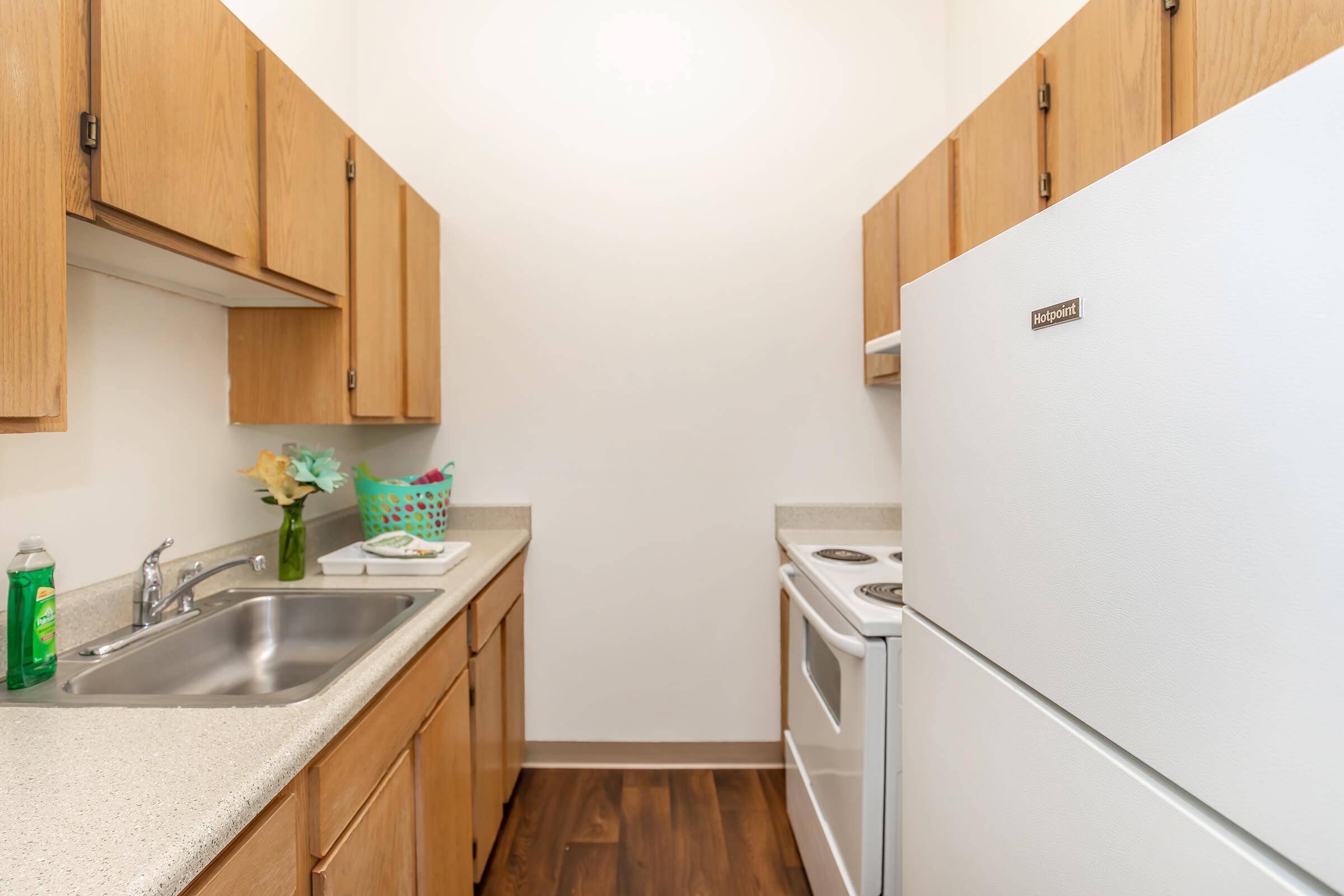 A small kitchen featuring light wooden cabinets, a white refrigerator, and a white stove. The countertop is light-colored with a sink, and there's a small floral arrangement and a colorful basket on it. The flooring is dark wood. The walls are painted a neutral color, providing a bright and spacious feel.