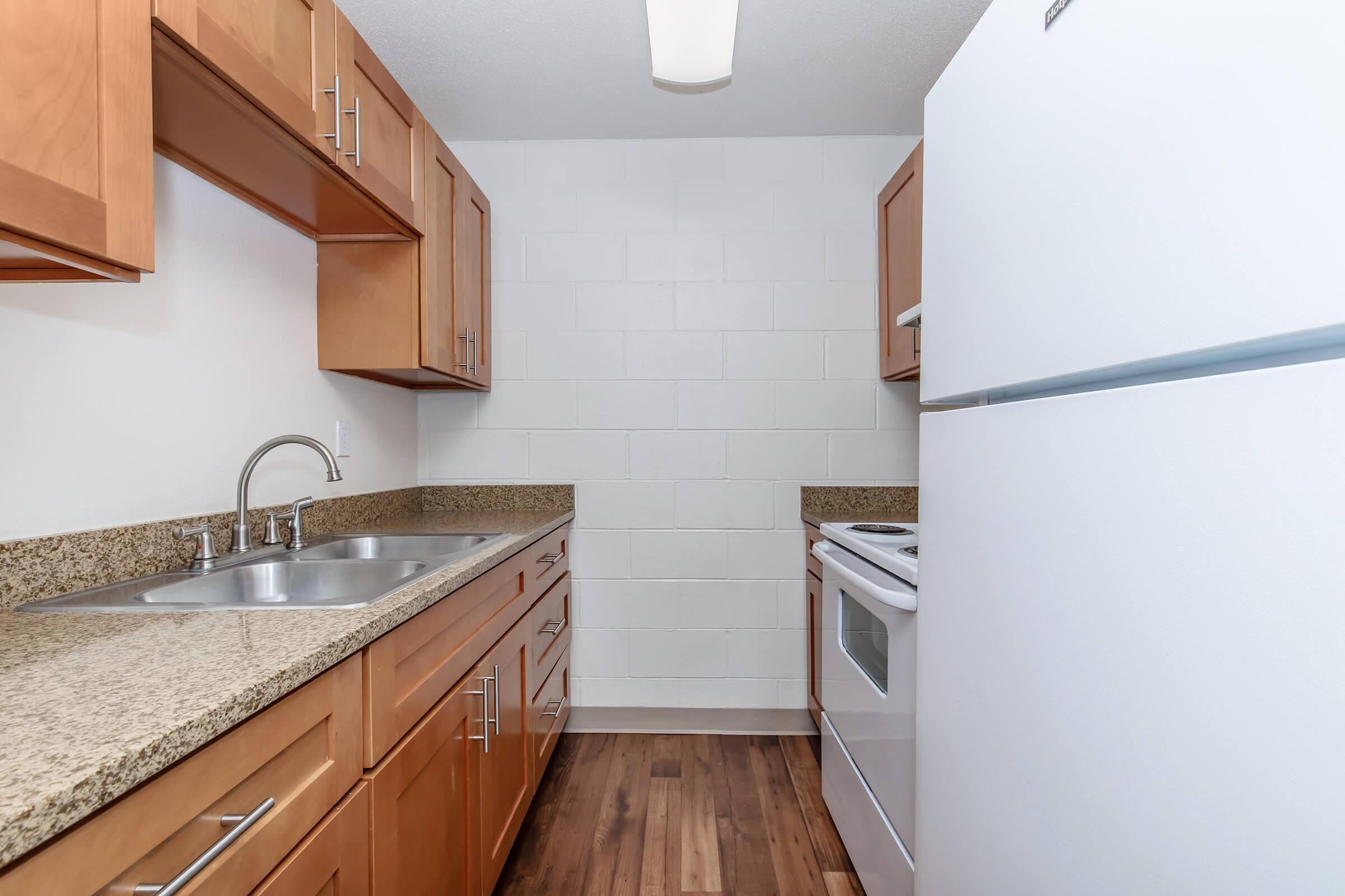 A bright kitchen featuring light wood cabinetry, a double sink with a modern faucet, and granite countertops. There is a white refrigerator and an electric stove, along with a tile backsplash and wooden flooring, creating a clean and inviting space. The walls are painted in a light color.