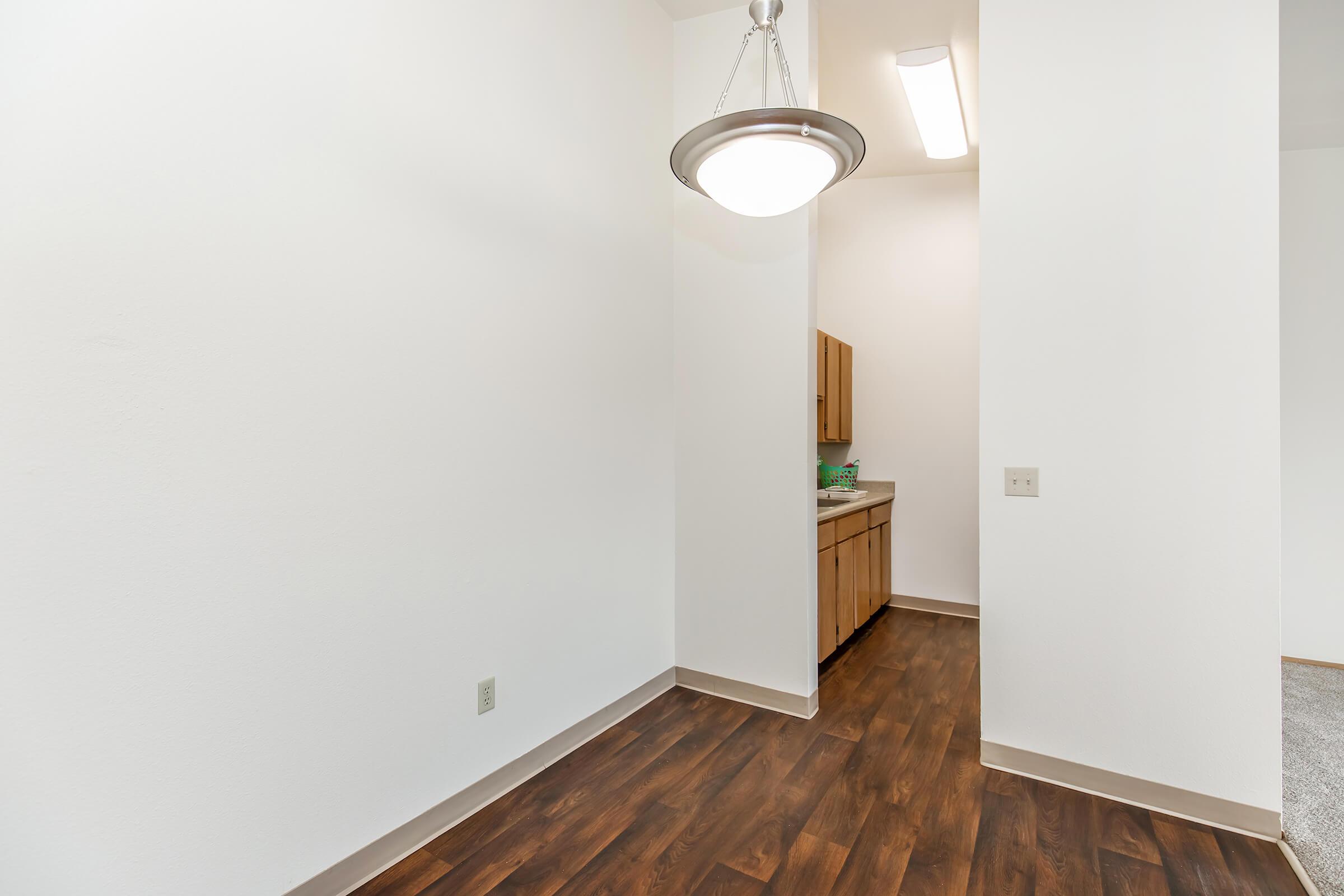 A well-lit kitchen area with white walls and dark wood laminate flooring. The space features a small kitchen counter with wooden cabinets and a sink. A pendant light hangs from the ceiling, illuminating the room. The area is clean and minimally furnished, showcasing an inviting atmosphere.