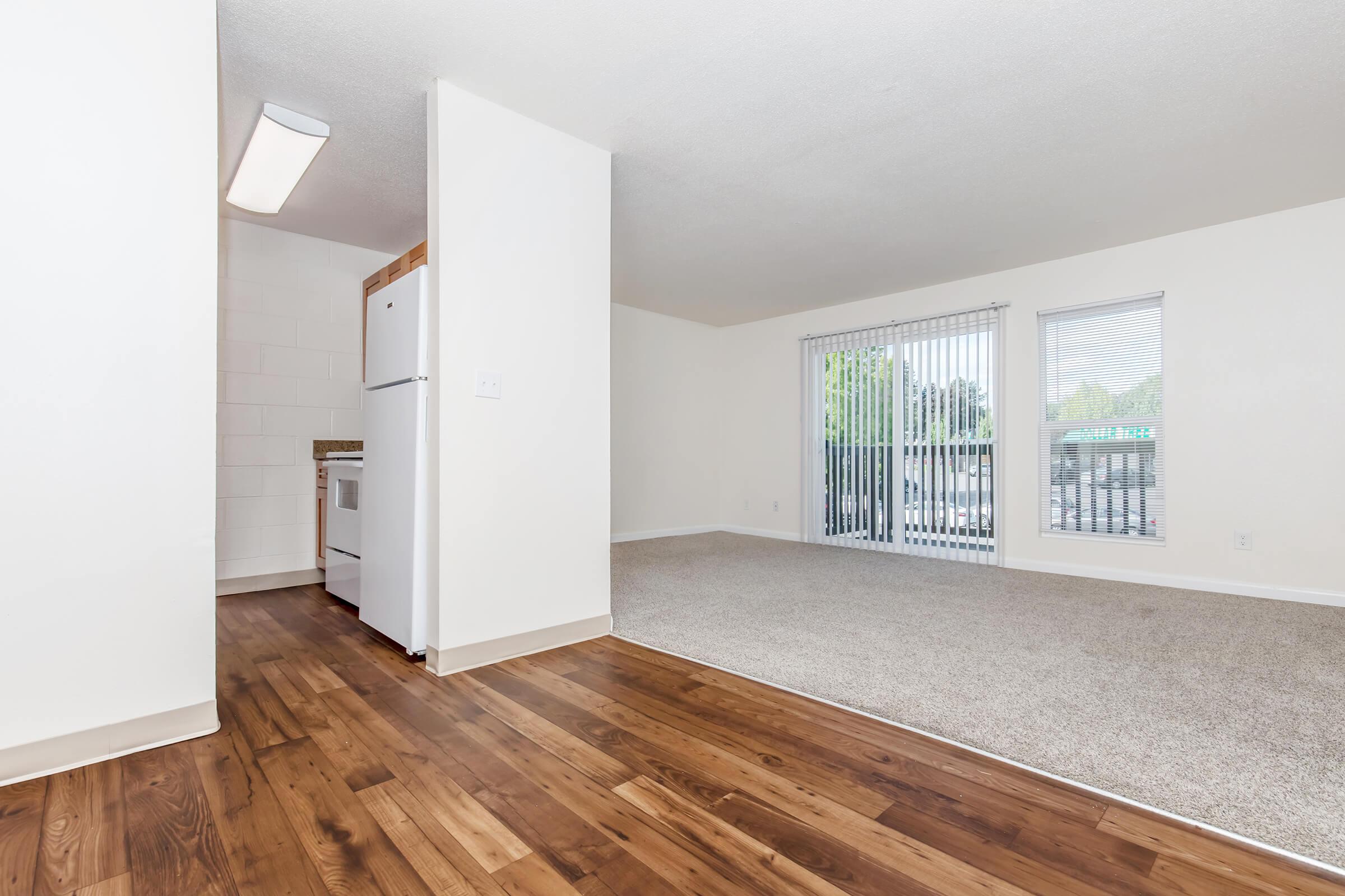 A spacious, well-lit living area with light-colored walls and a large window. The flooring features a combination of carpet and wood, connecting to a kitchen area visible on the left. A sliding glass door leads to an outdoor space, enhancing the open feel of the room.
