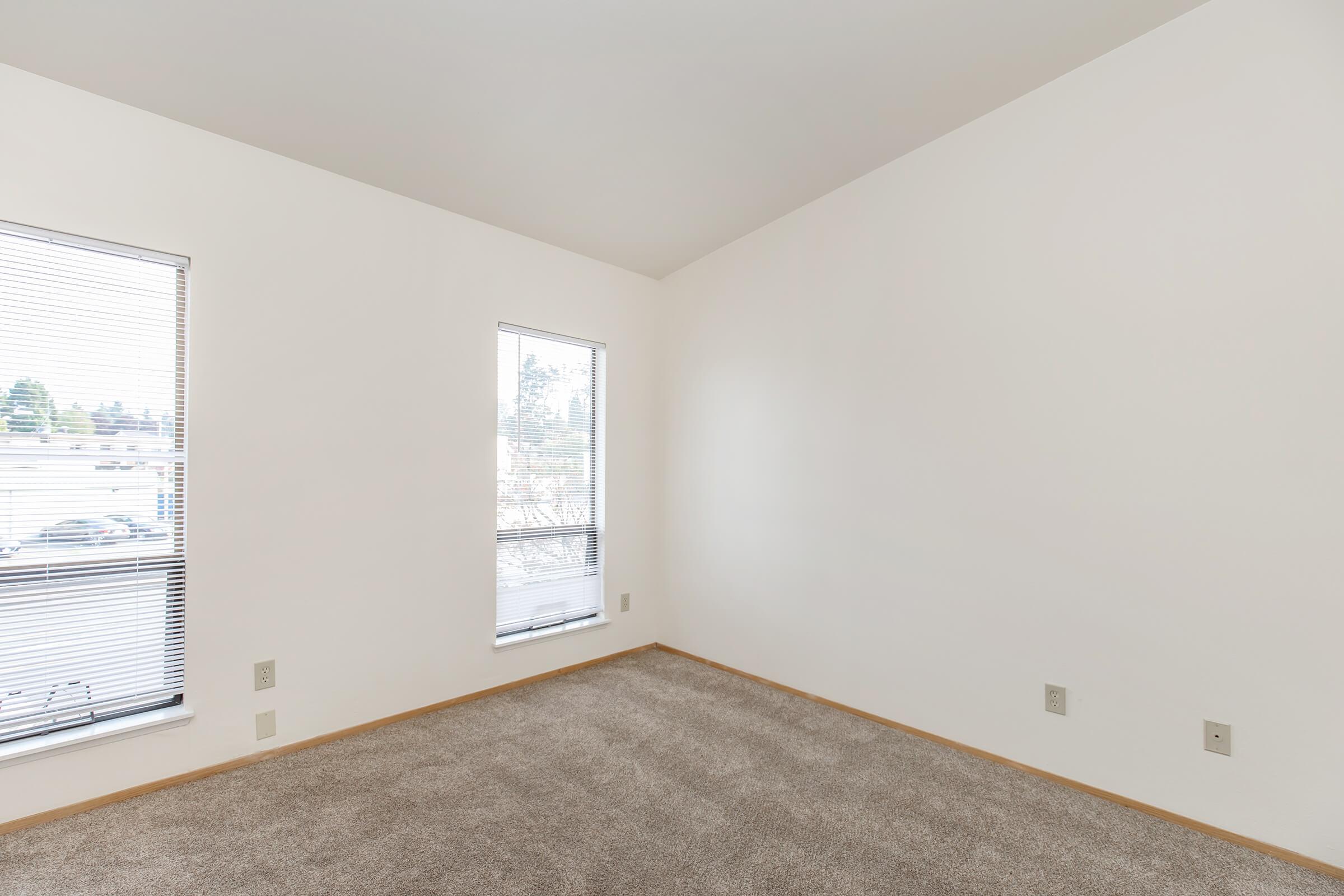 Empty room with light beige walls and carpeting. Two windows with blinds allow natural light to enter. The corner of the room is visible, showcasing the clean and minimalist aesthetic. A few electrical outlets are present on the walls, but no furniture or decorations are included.