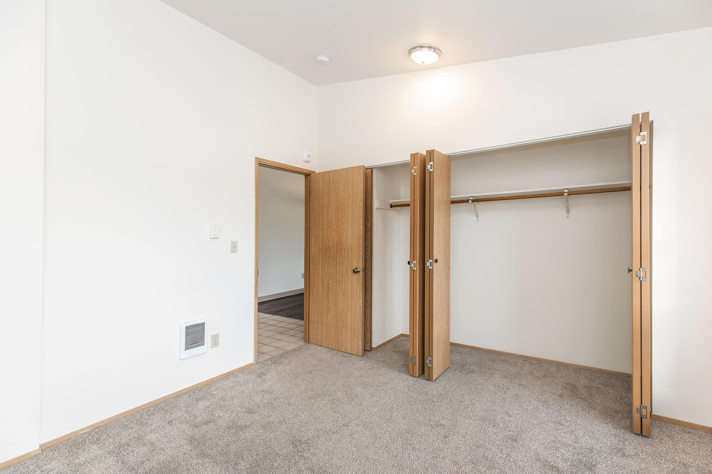 A newly renovated room featuring light-colored walls and plush beige carpet. It includes a spacious closet with double doors, providing ample storage space. A soft overhead light illuminates the area, and a doorway leads to another room in the background.