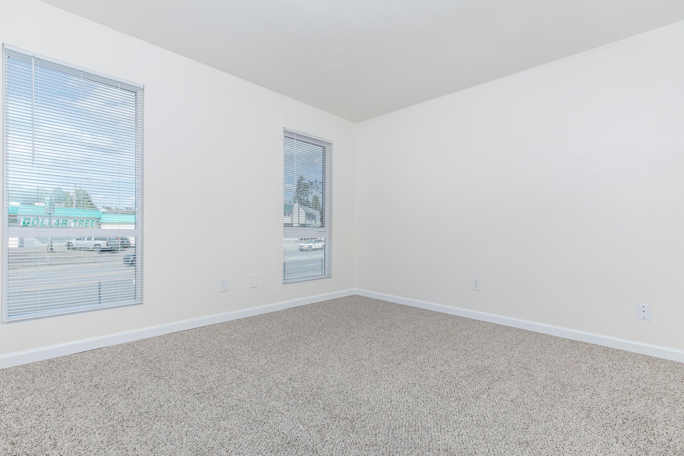 Empty room with light beige walls and a carpeted floor. There are two windows with blinds allowing natural light to enter. The space is bright and uncluttered, ideal for personalized decoration or use as an office or living area.
