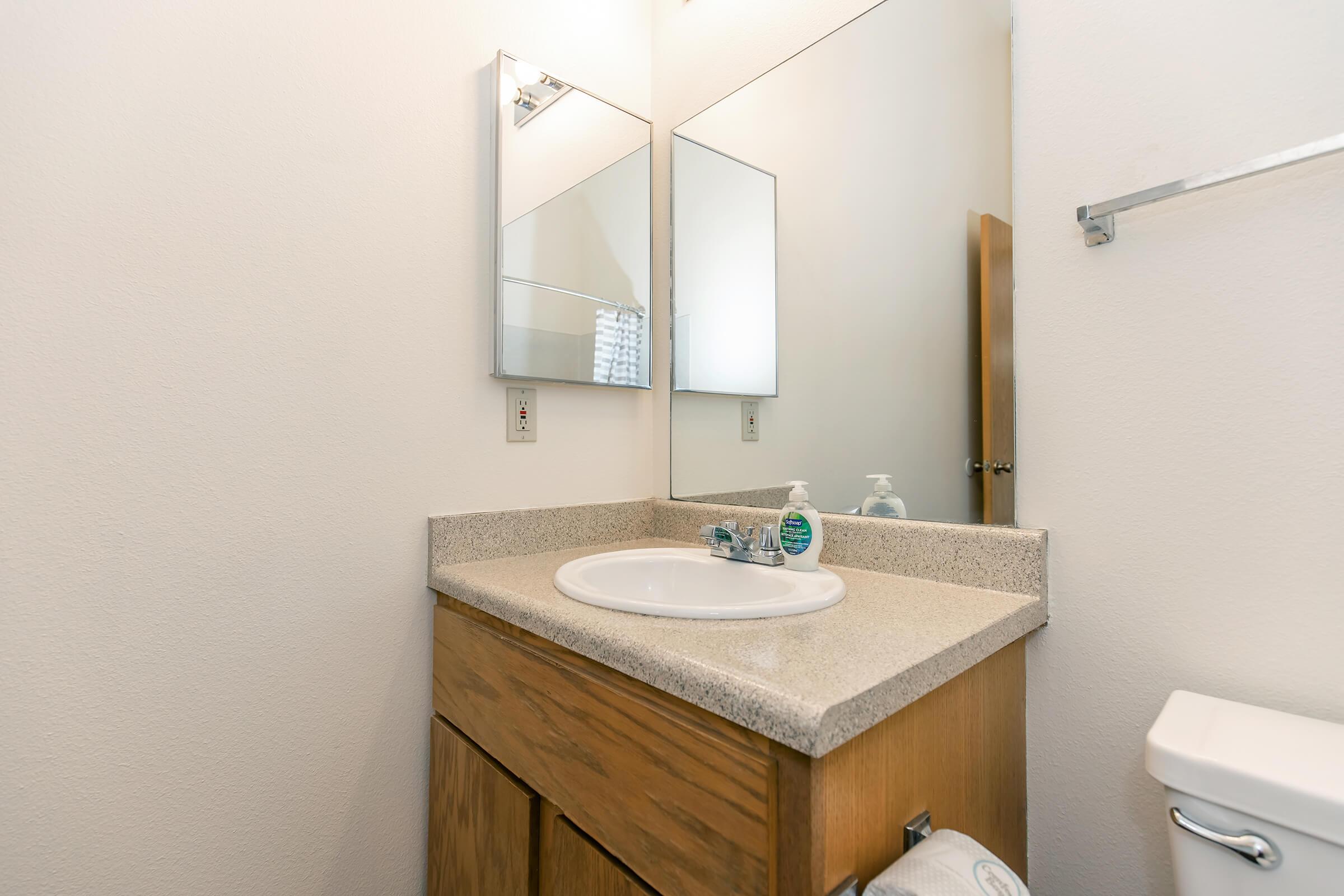 A clean, modern bathroom featuring a light-colored countertop with a sink, two mirrors above the sink, and a wooden cabinet below. The walls are painted white, and there is a toilet to the right. Hand soap and sanitizer are visible near the sink. Natural light may come through a window not shown in the image.