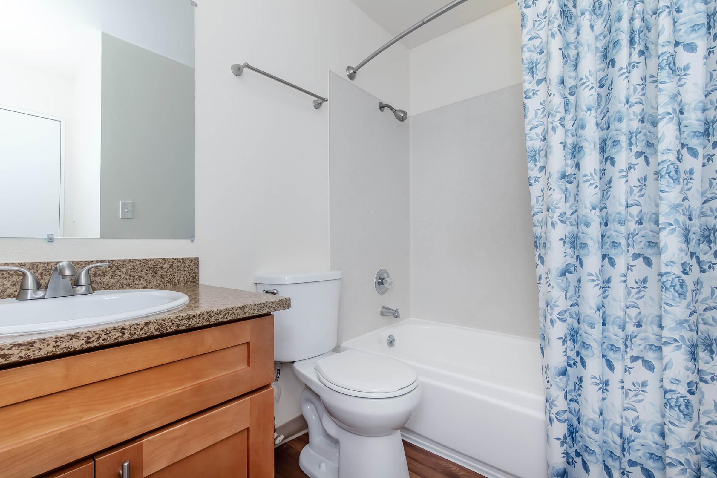 A clean, modern bathroom featuring a bathtub with a shower curtain adorned with blue floral patterns. There's a sink with a granite countertop and wooden cabinetry. A mirror is mounted on the wall, reflecting the well-lit space. The floor has a wood-like finish, adding warmth to the design.