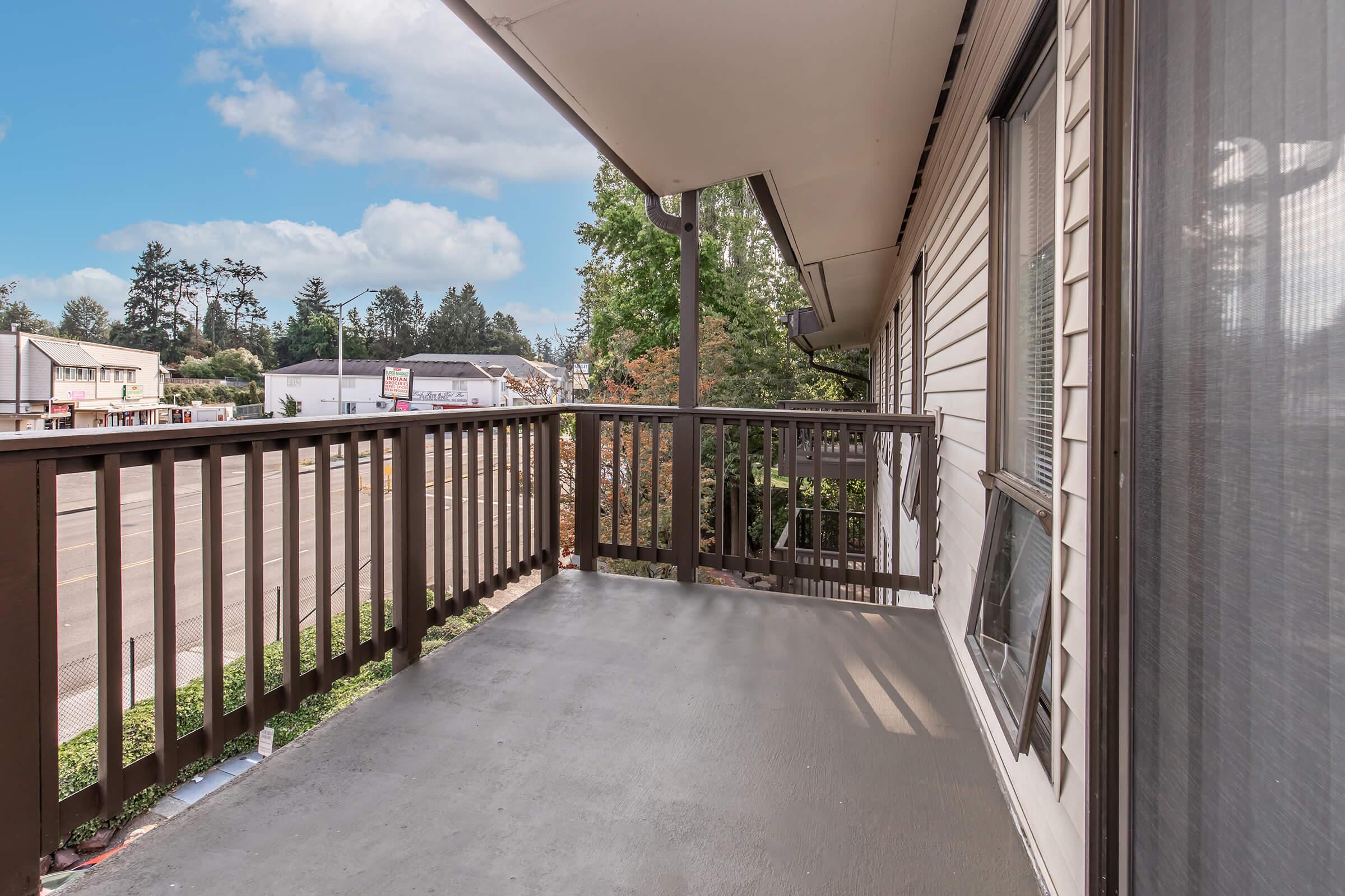 A spacious balcony with a wooden railing, overlooking a parking lot and nearby buildings. The scene is framed by trees and a blue sky with scattered clouds, creating a bright and inviting atmosphere. The floor is clean and well-maintained, enhancing the open feel of the outdoor space.