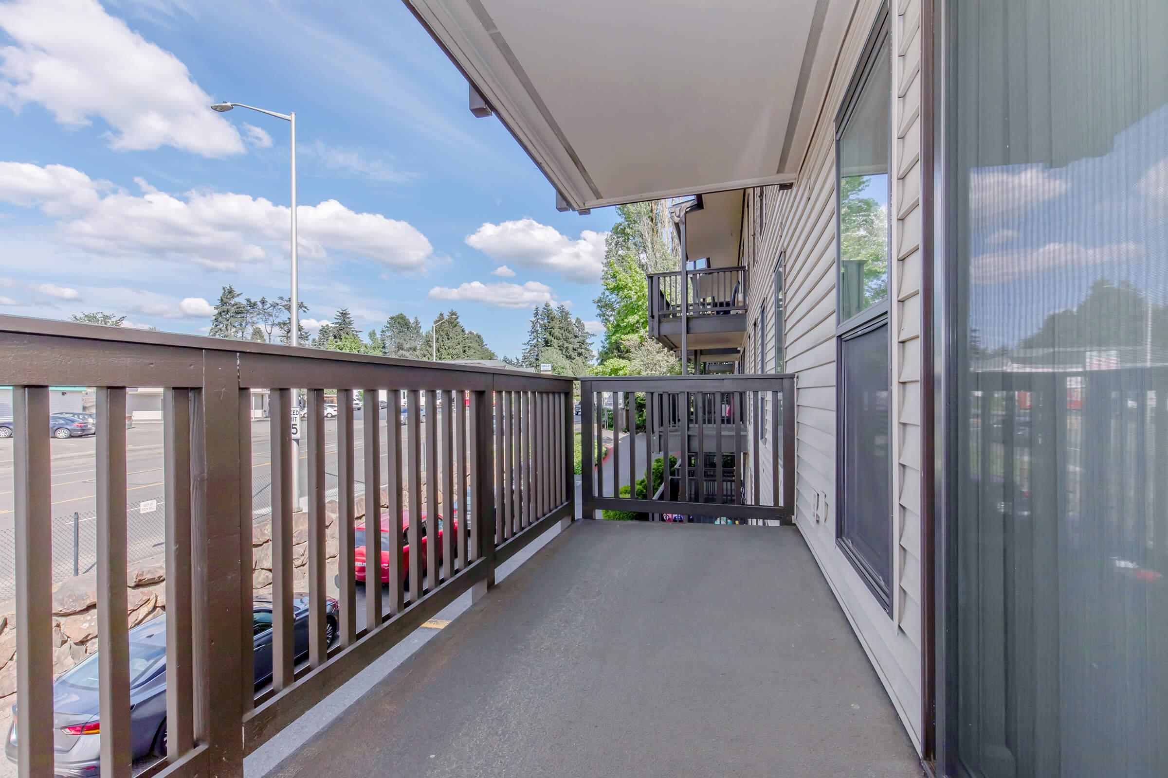 A view from a balcony featuring wooden railing, a concrete floor, and a clear sky with fluffy clouds. In the background, there are green trees and parked cars. The scene conveys a sense of openness and outdoor space.