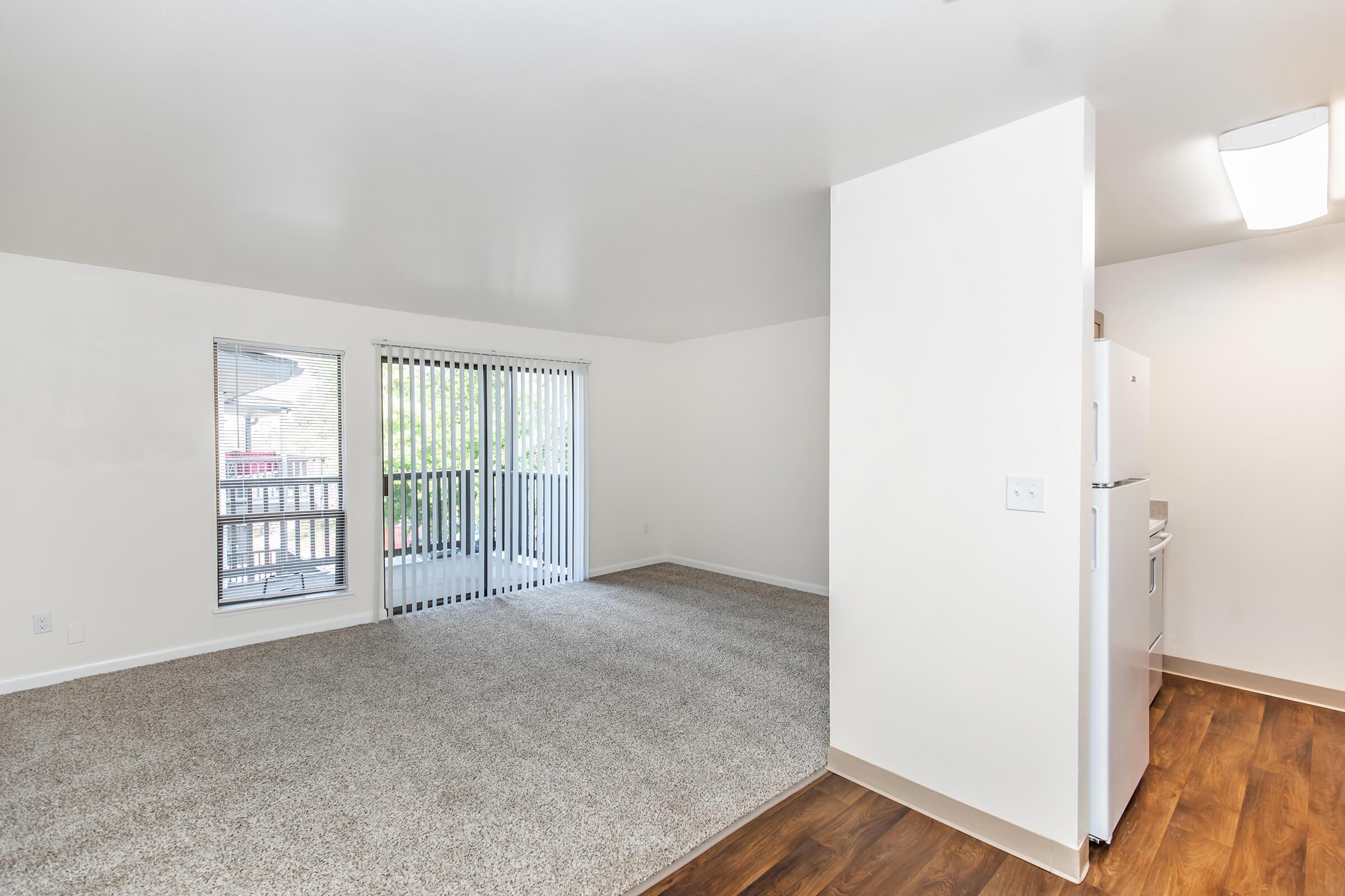 A spacious, light-filled living area with beige carpeting and white walls. Large windows with vertical blinds provide natural light and a view of a balcony. To the right, a small kitchen area is visible, featuring white appliances and cabinetry. The overall feel is modern and inviting, perfect for a comfortable living space.