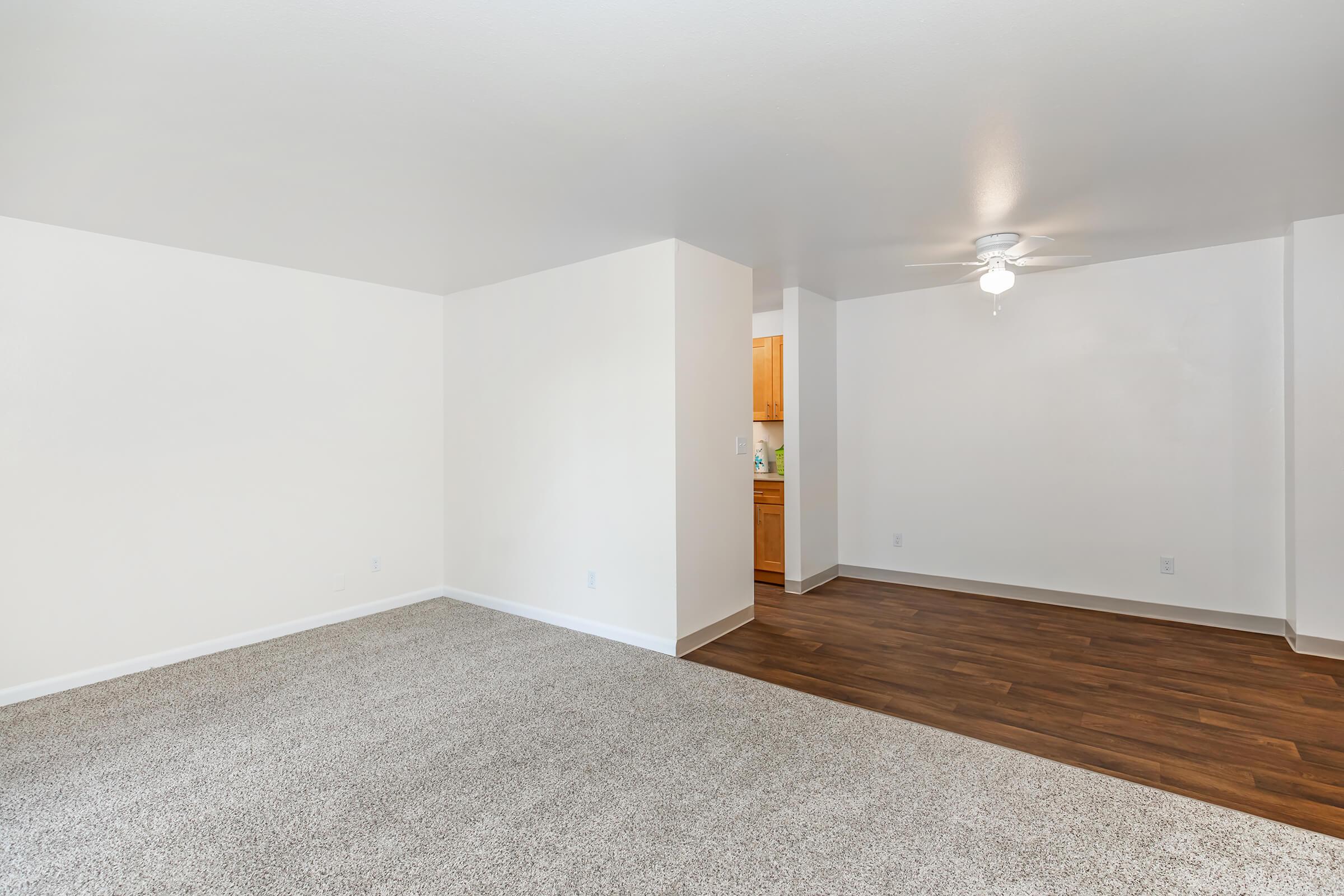 A spacious, empty living area with light-colored walls and carpeting, featuring a ceiling fan and a view into an adjacent kitchen area with wooden cabinetry. The room has ample natural light, creating a warm and inviting atmosphere.