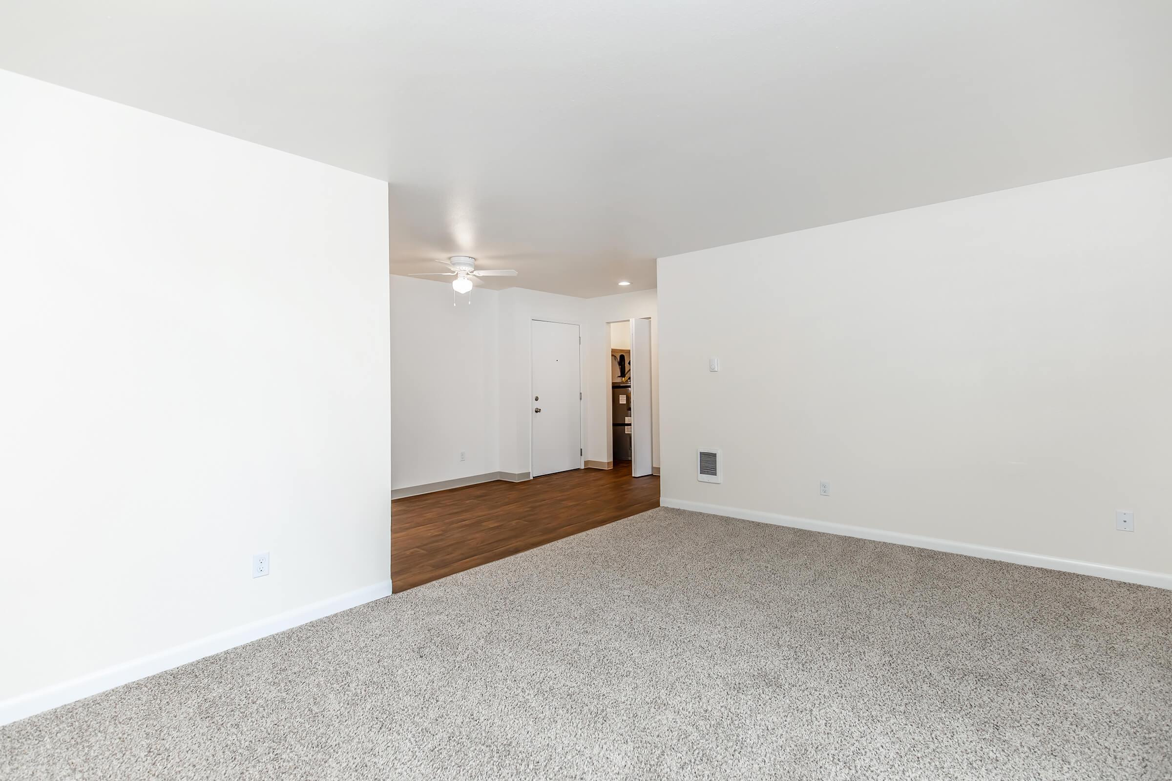 Spacious living area featuring light-colored walls, a ceiling fan, and soft carpet. To the right, there is a doorway leading to a kitchen area. The overall ambiance is bright and inviting, with ample room for furniture and decor.