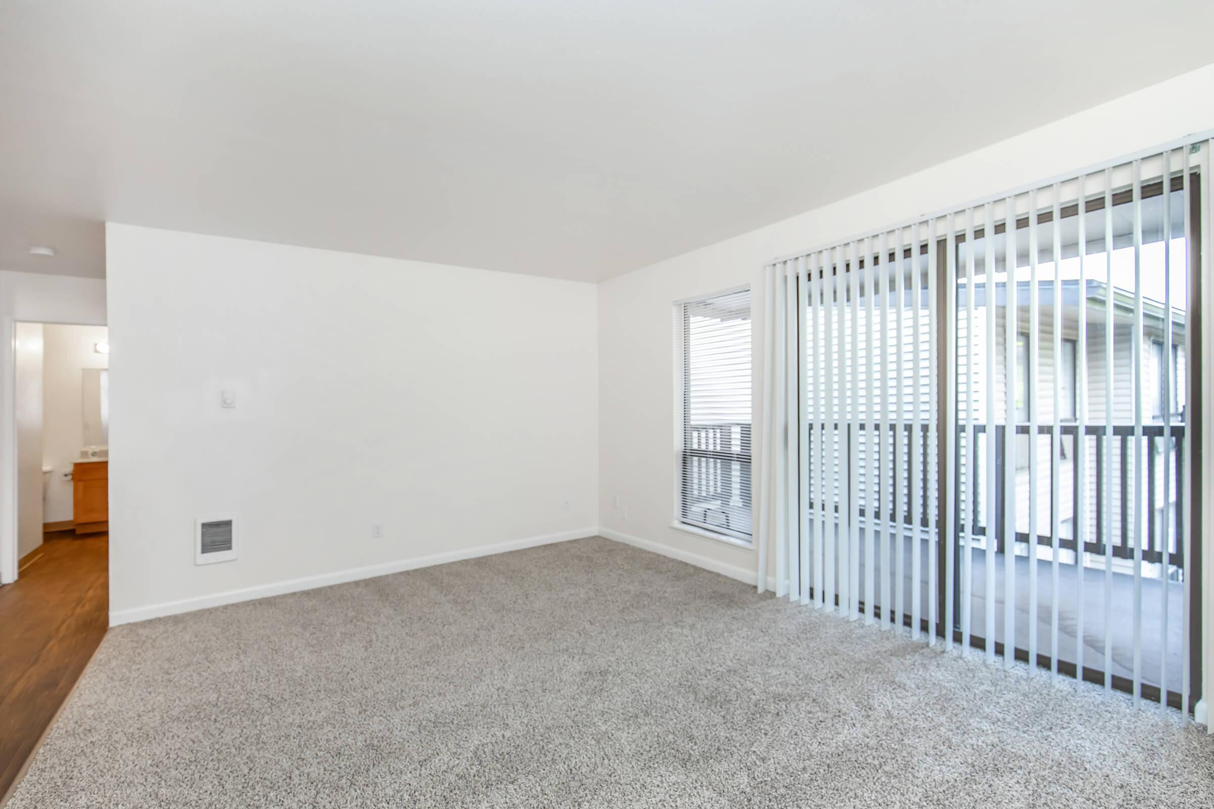 Empty living room featuring beige carpet and white walls. Large sliding glass doors with vertical blinds lead to a balcony. A doorway on the left connects to a bathroom area. Natural light brightens the space, creating a welcoming atmosphere.