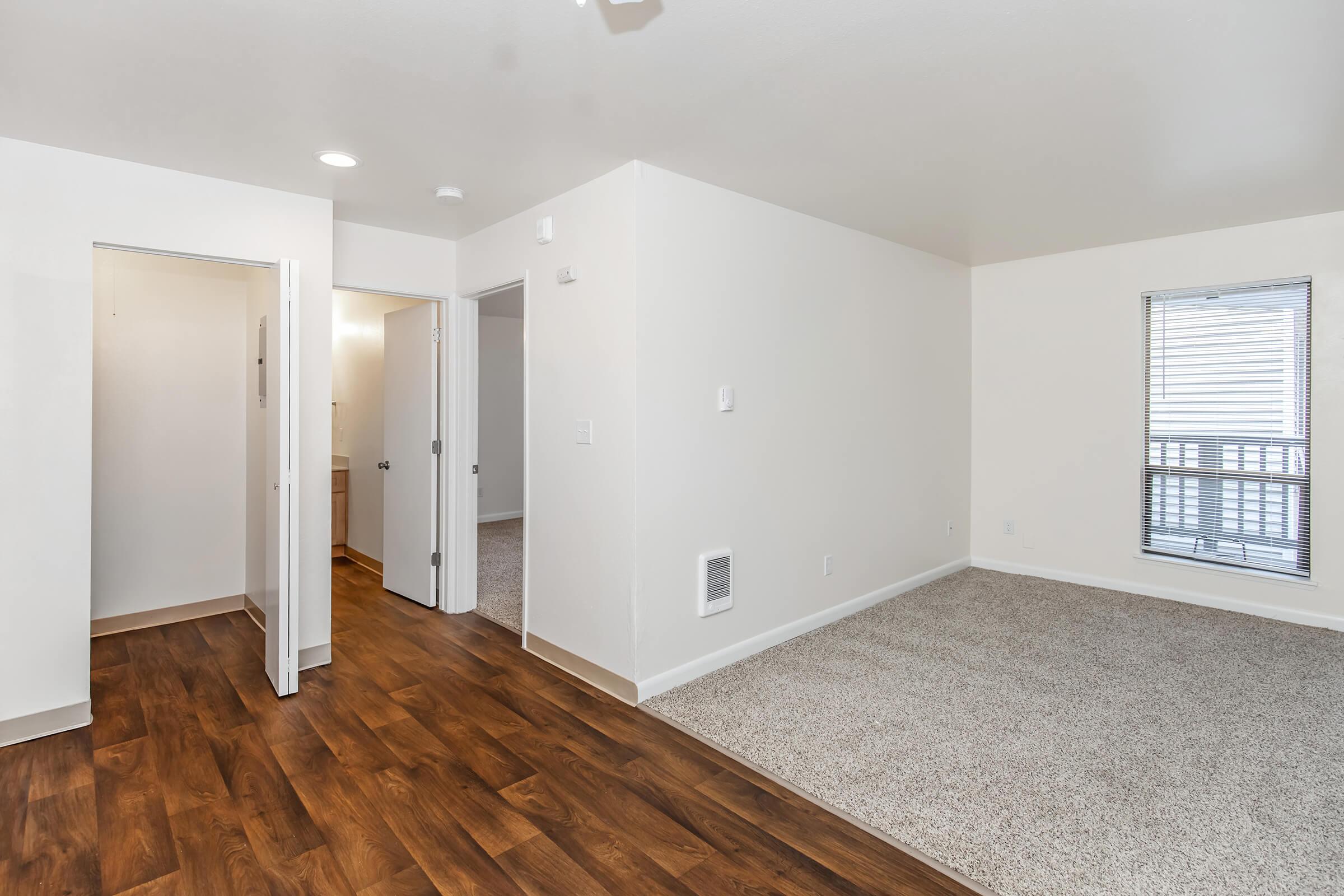 Interior view of a spacious, well-lit room featuring beige walls, wooden flooring, and carpeted areas. The layout includes open spaces with doors leading to other areas, such as a bathroom. A window provides natural light, contributing to a bright and inviting atmosphere.