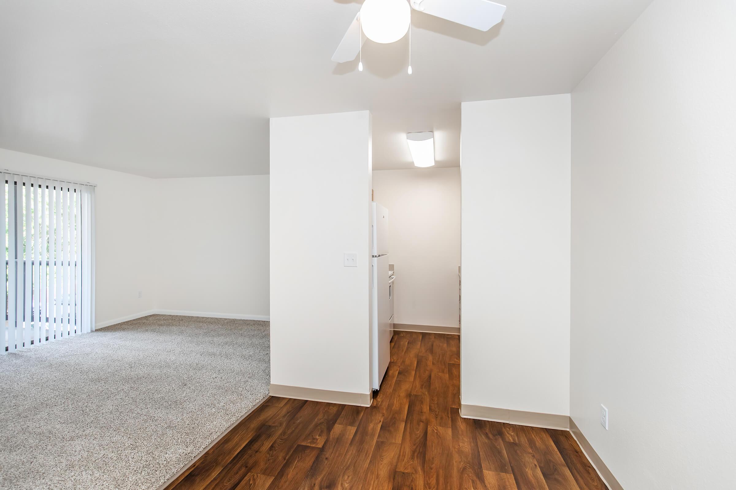 A bright, spacious interior of an apartment featuring light-colored walls, a ceiling fan, and carpeted flooring. On one side, there is an open living area with sliding glass doors leading to a balcony, while on the other, a white kitchen area is visible with a refrigerator and modern fixtures.
