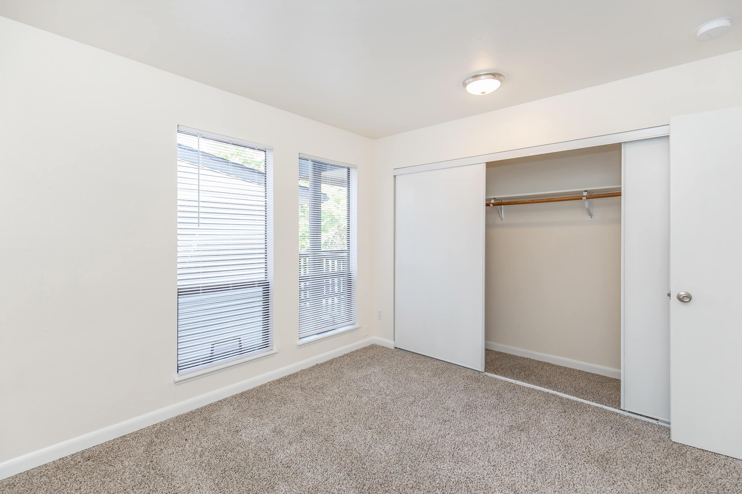 A well-lit, empty bedroom with light-colored walls and carpeted flooring. Two large windows with horizontal blinds allow natural light in. There is a sliding closet door on the right, showcasing a simple closet with a hanging rod. The overall space appears clean and ready for furnishing.