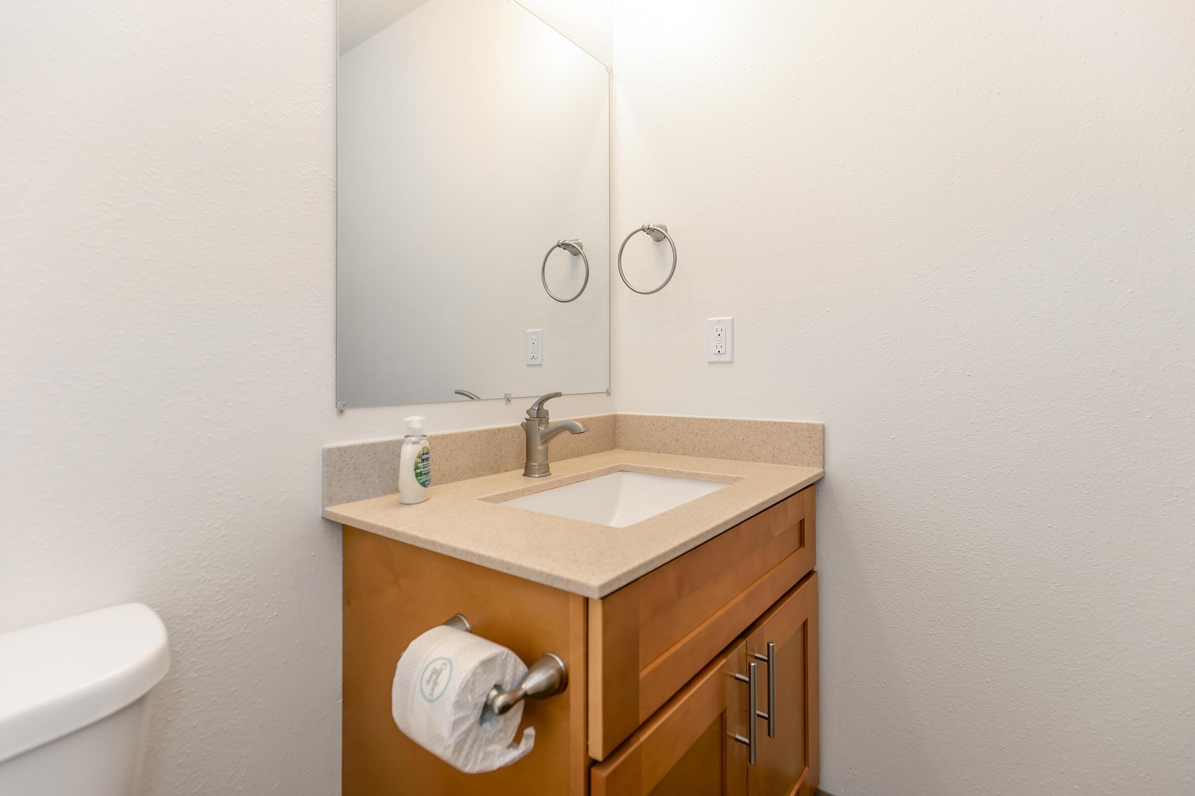 A clean, modern bathroom with a light-colored wall, a small wooden vanity featuring a sink and faucet, a large mirror above the sink, a paper towel holder on the side, and a toilet visible on the left side. The space appears bright and well-maintained.