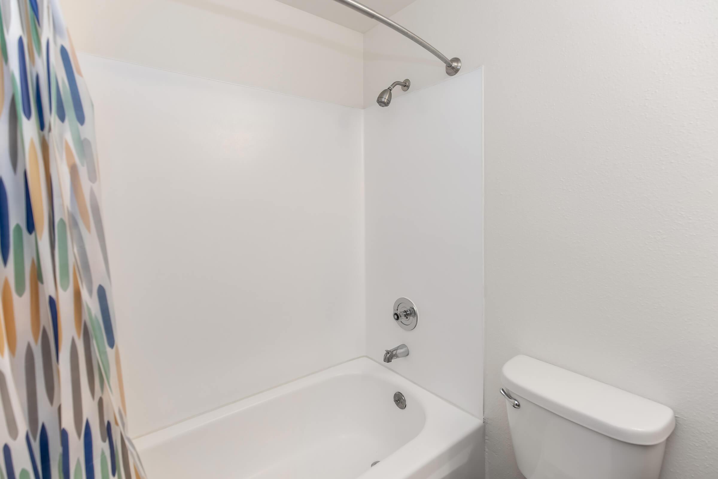 A clean, white bathroom featuring a bathtub with a showerhead, a shower curtain with colorful polka dots, and a toilet nearby. The walls are white, and the overall space is well-lit and minimalistic.