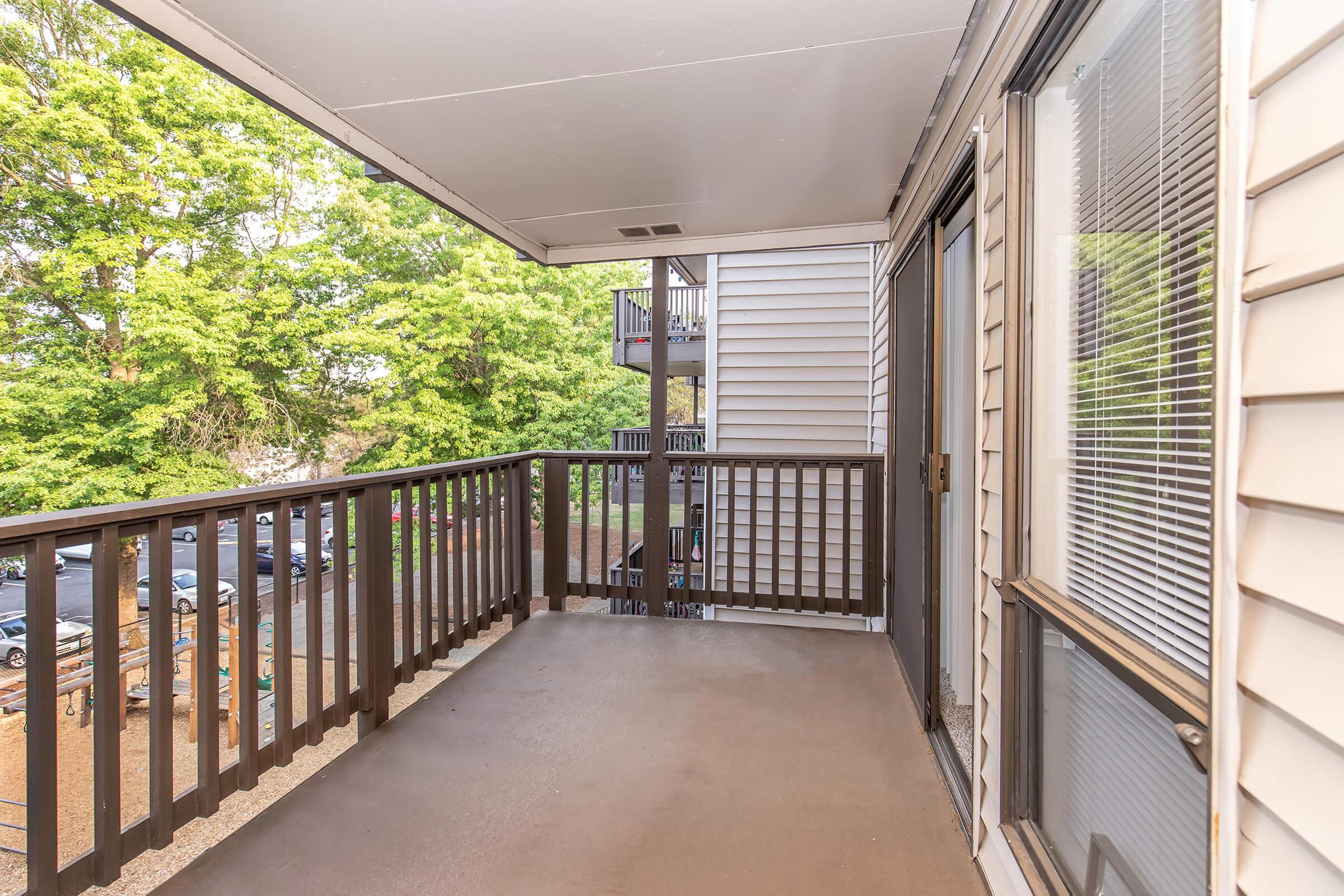 A view of a balcony with wooden railings overlooking green trees. The balcony features a concrete floor and has a door leading into an interior space. In the background, there are glimpses of parked cars and an adjacent building. Natural light brightens the area, creating a welcoming atmosphere.