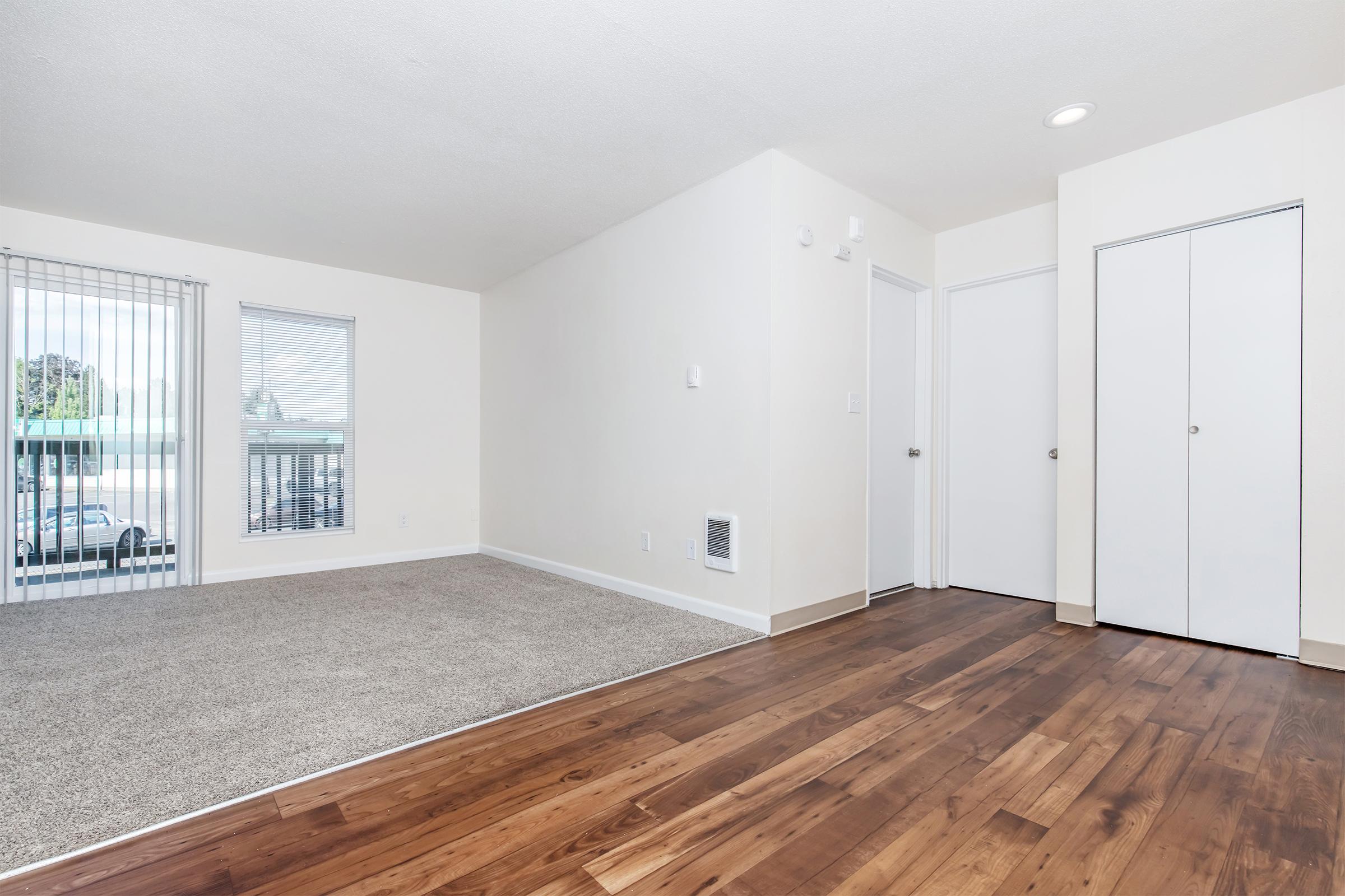A spacious living area featuring beige walls, light brown wooden flooring, and plush carpet. There's a door leading to an outside area and a wall of windows with vertical blinds. A closet is visible to the right, and a white vent is seen on the wall, enhancing the room's inviting atmosphere.