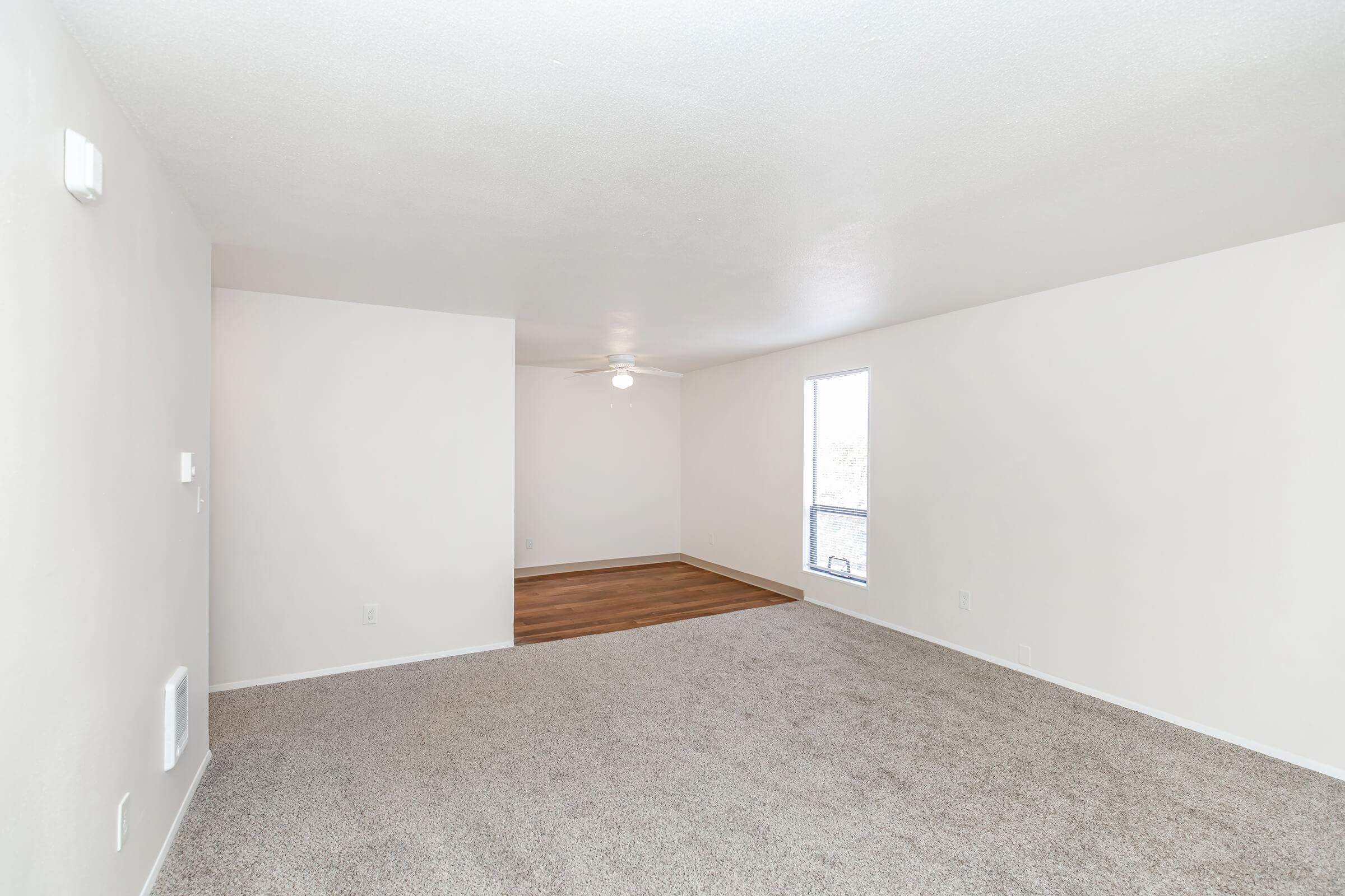 A spacious, empty living room with light-colored walls and carpet. A ceiling fan is visible, and there is a large window allowing natural light to fill the space. The flooring transitions to hardwood in the adjacent area, creating a cozy and inviting atmosphere.