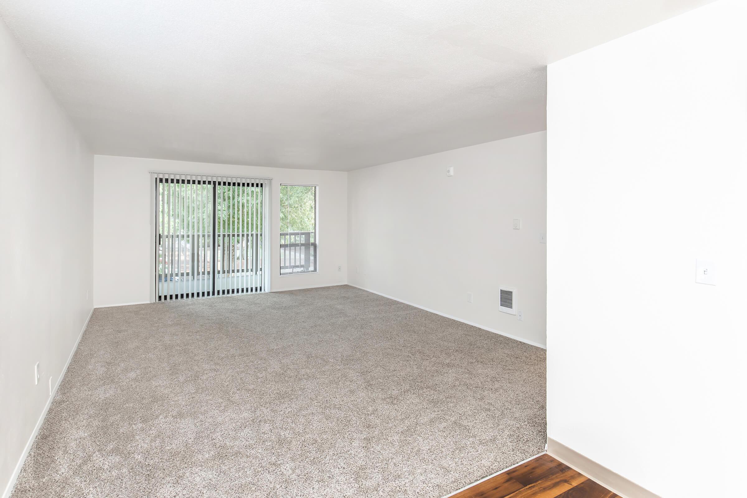 A spacious and empty living room with light beige carpet, white walls, and sliding glass doors leading to a balcony. Natural light filters in through the windows, creating a bright atmosphere. The room is unfurnished, providing a blank canvas for decorating.