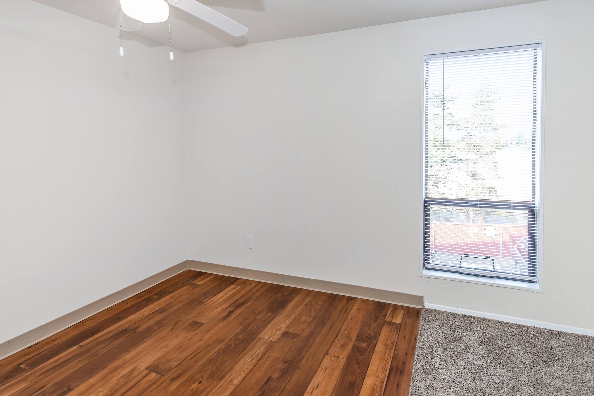 A spacious, empty room featuring light-colored walls, a ceiling fan, and a large window with blinds. The floor is a mix of polished wood and carpet, creating a modern and inviting atmosphere. Natural light is visible through the window, highlighting the room's layout.