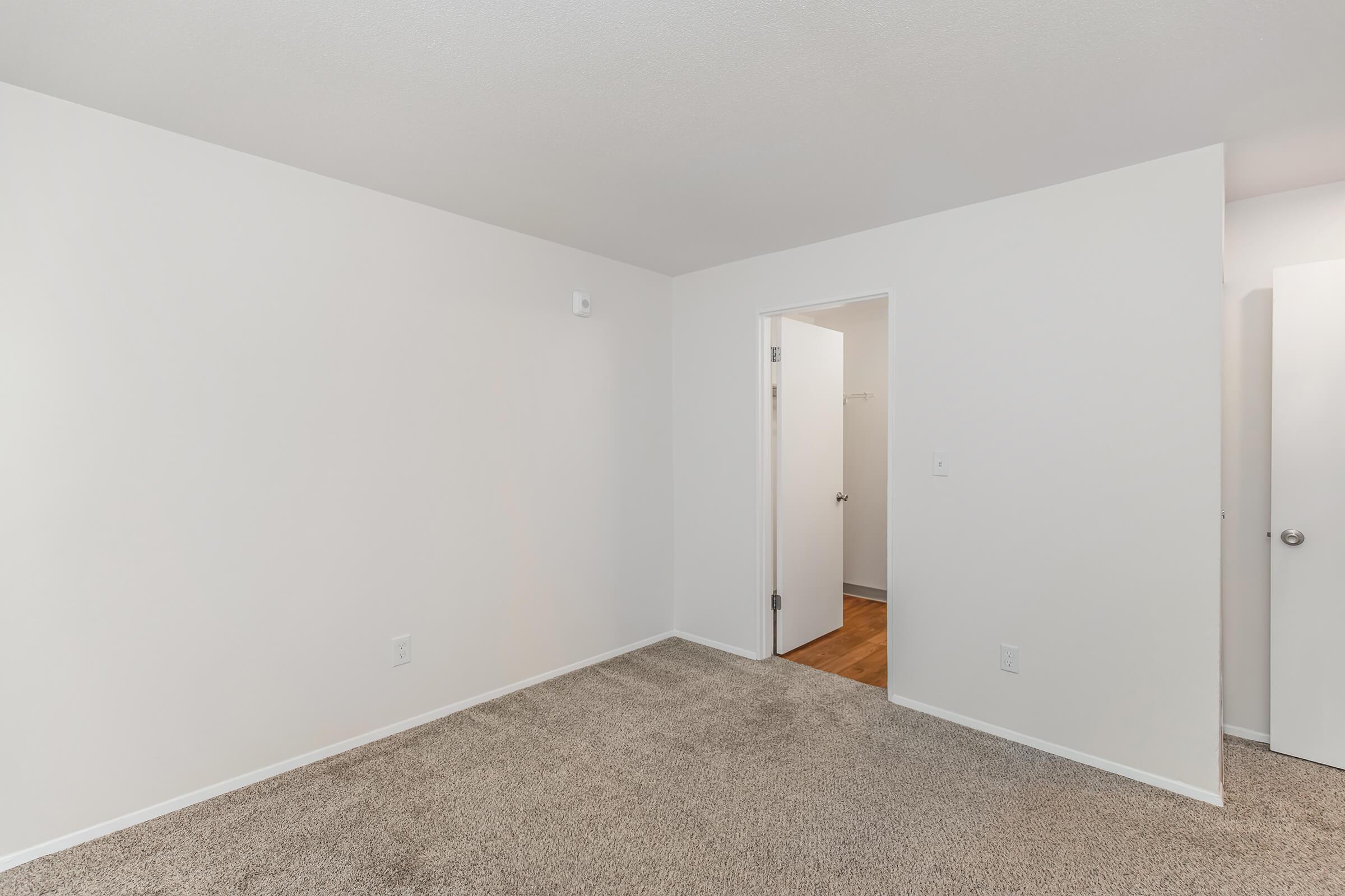 A spacious, empty room featuring light beige carpet and white walls. A door leads to another room, and a second door is partially visible on the right. The space is well-lit and minimalistic, ideal for various design possibilities.