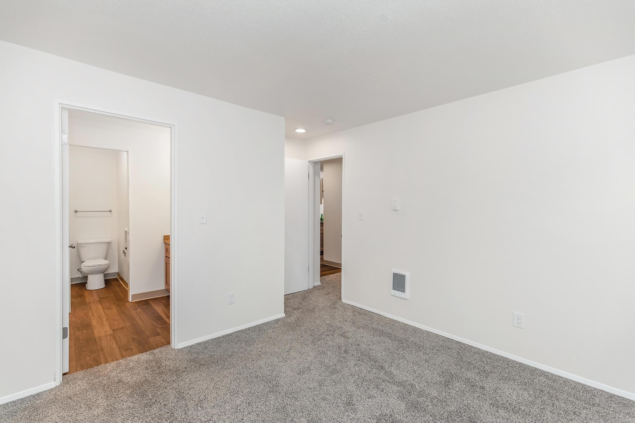 A spacious, light-filled interior of an apartment featuring beige carpet flooring. A doorway leads to a small bathroom with a toilet visible, and another door opens to a hallway. The walls are painted white, and there are minimal furnishings in the area.