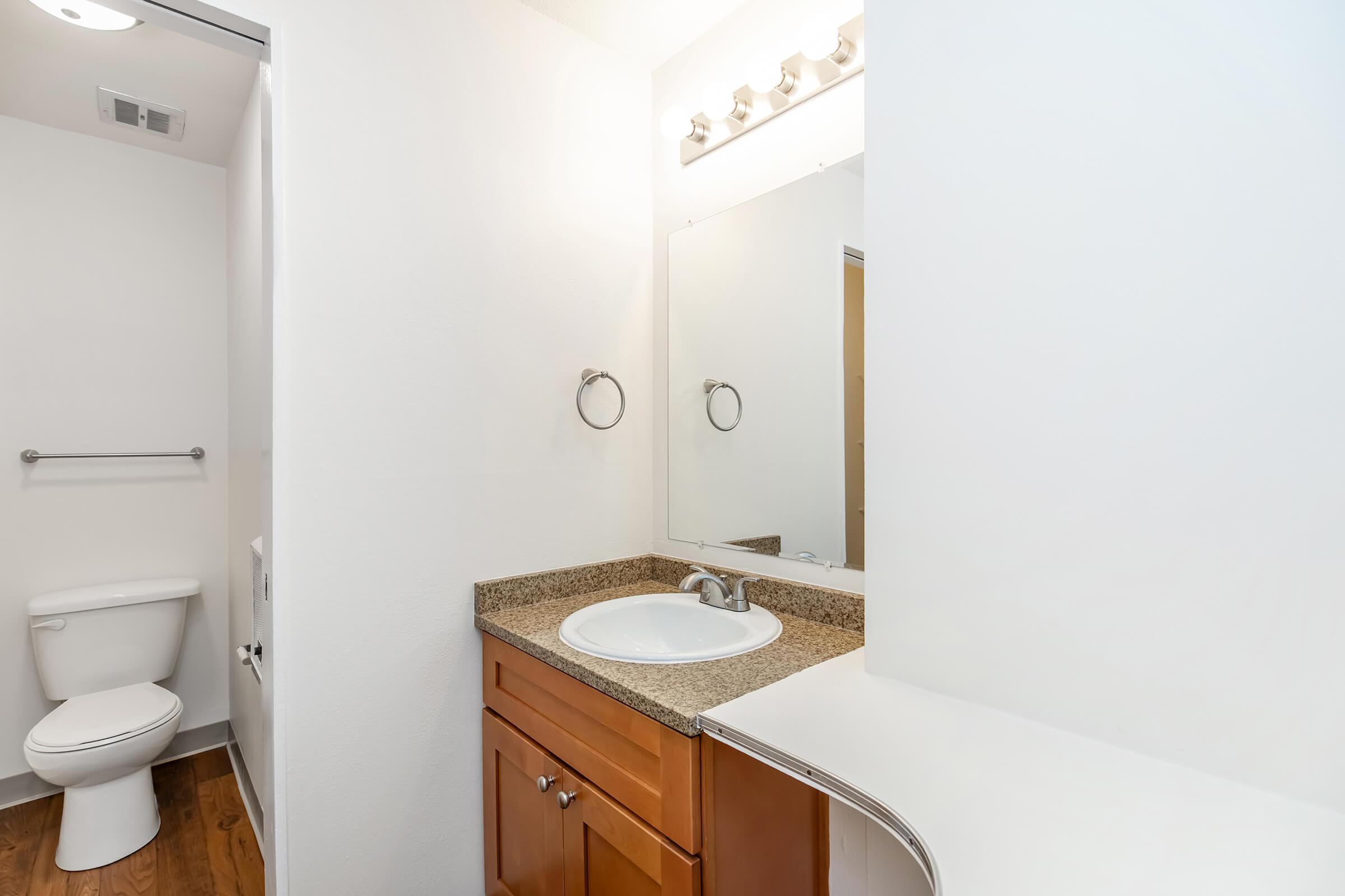 A bright bathroom featuring a white wall, a wooden floor, and a modern vanity with a round sink. Above the sink, there are five light fixtures, and a large mirror is mounted on the wall. A toilet is visible in the background, with a towel rack beside it.