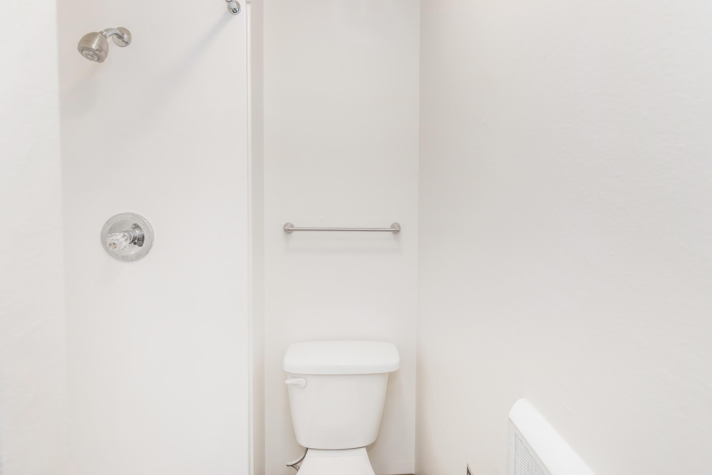 A clean and minimalistic bathroom featuring a white toilet and a shower with a simple door. The walls are painted white, creating a bright and modern look. A chrome towel bar is mounted on the wall beside the shower, enhancing the functionality of the space.