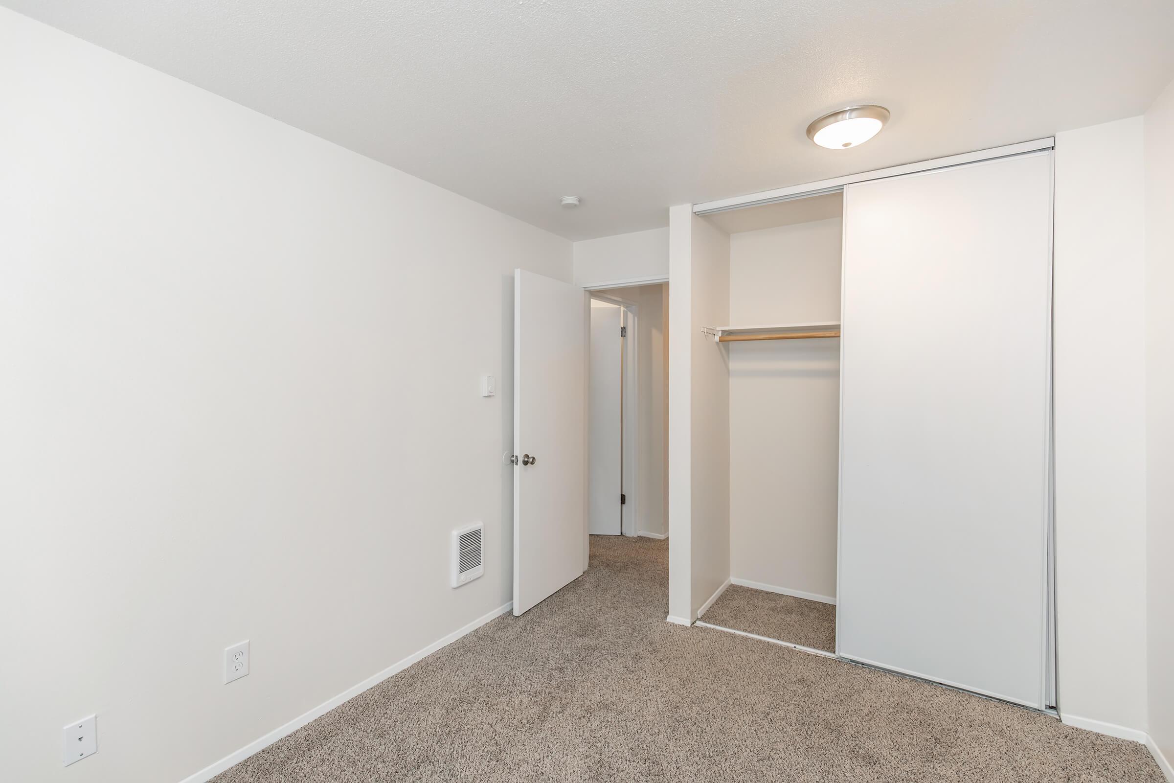 A view of an empty room featuring beige carpet, plain white walls, and a ceiling light fixture. There are two doors: one leading to another room and the other to a closet with a hanging rod. The space appears clean and well-lit, offering a blank canvas for decoration.
