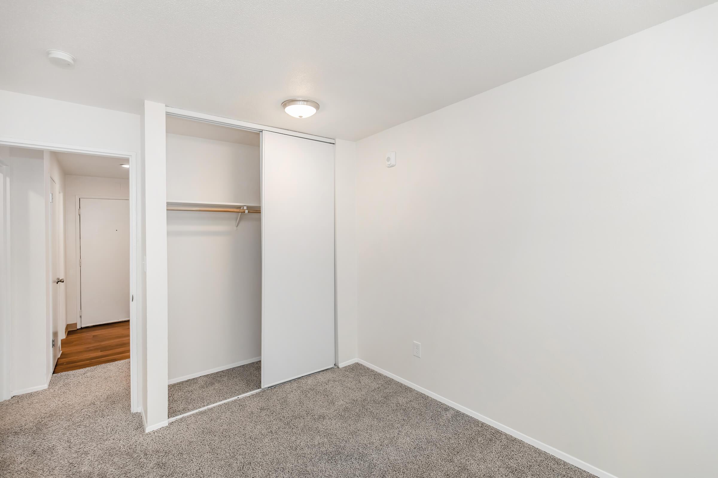 Empty room with light-colored walls and carpeted flooring. A sliding door closet with a shelf is visible on the left side, and there is a door leading to another room. Natural light from an overhead light fixture illuminates the space, creating a clean and minimalistic atmosphere.
