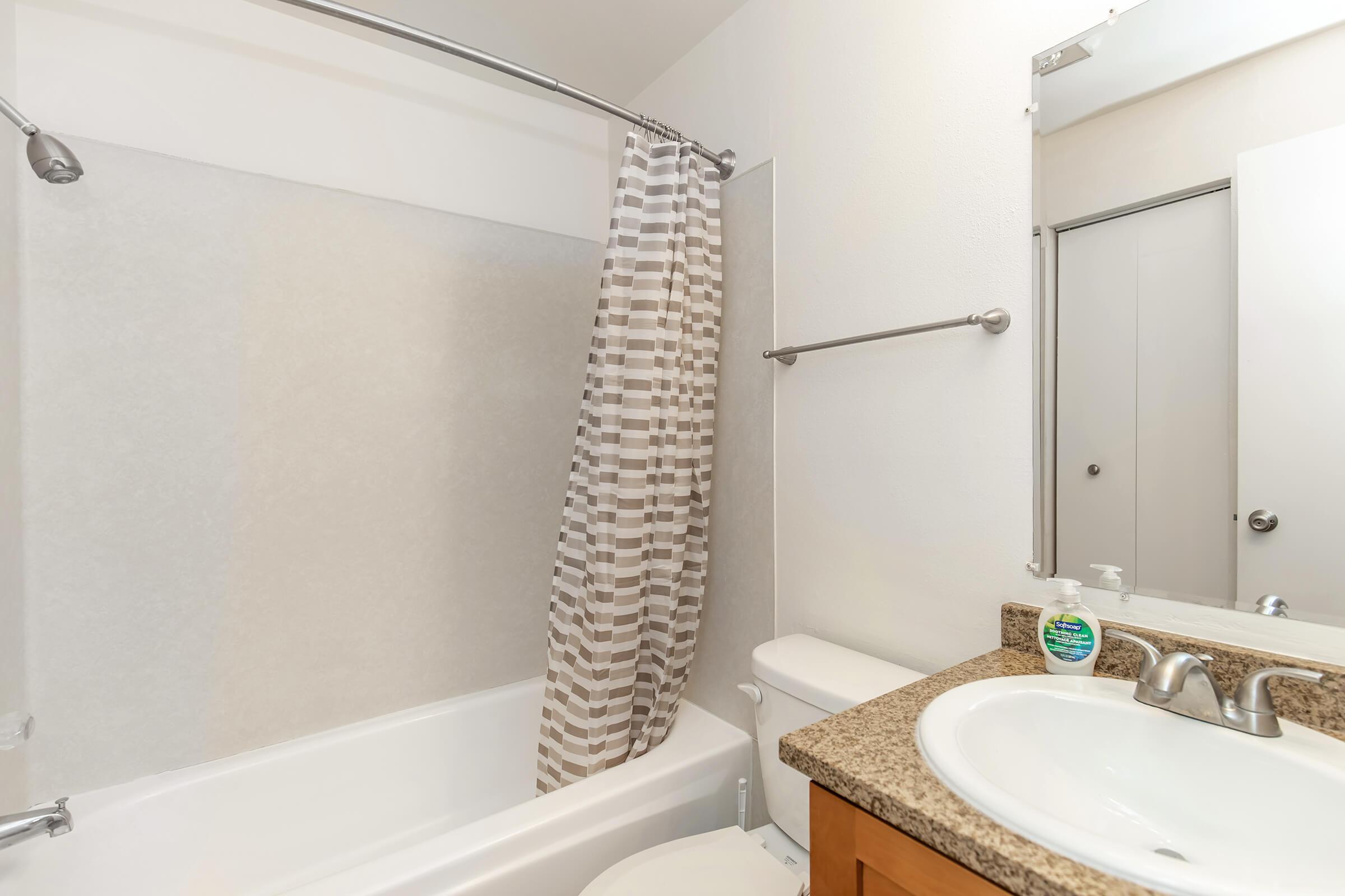 A clean and simple bathroom featuring a shower/bathtub combination with a striped shower curtain, a granite countertop with a soap dispenser, and a toilet. The walls are light-colored, and there is a mirror above the sink.