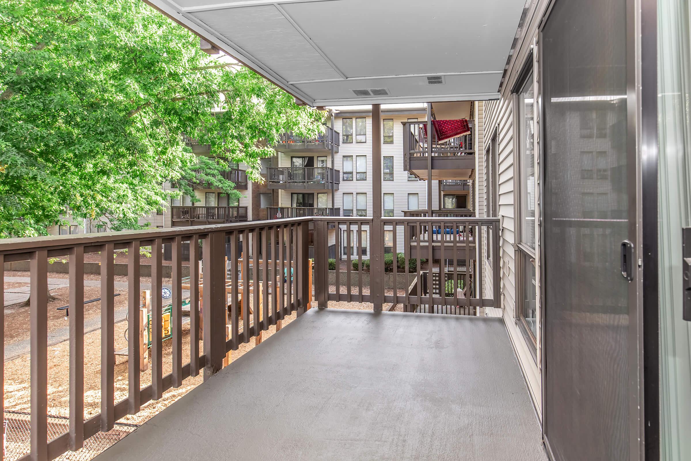 View of a spacious balcony with a wooden railing overlooking a landscaped courtyard. In the background, multiple apartment buildings are visible, surrounded by trees. The floor is finished with a smooth surface, creating a welcoming outdoor space. Bright natural light enhances the inviting atmosphere.