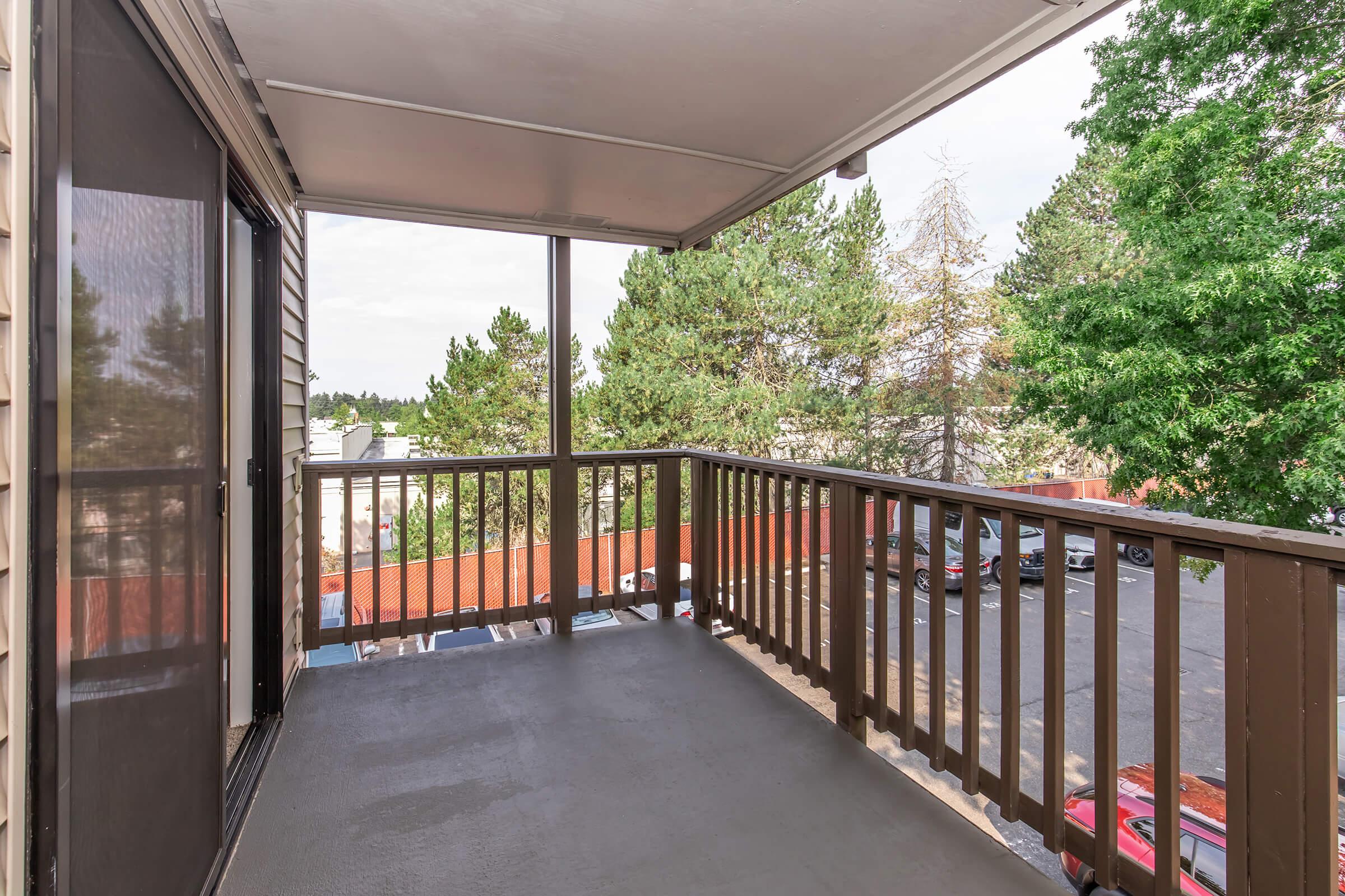 A view from a balcony with a wooden railing, overlooking a parking lot and trees. The floor is gray, and there is a screen door on one side. Soft sunlight brightens the space, creating a welcoming outdoor area.