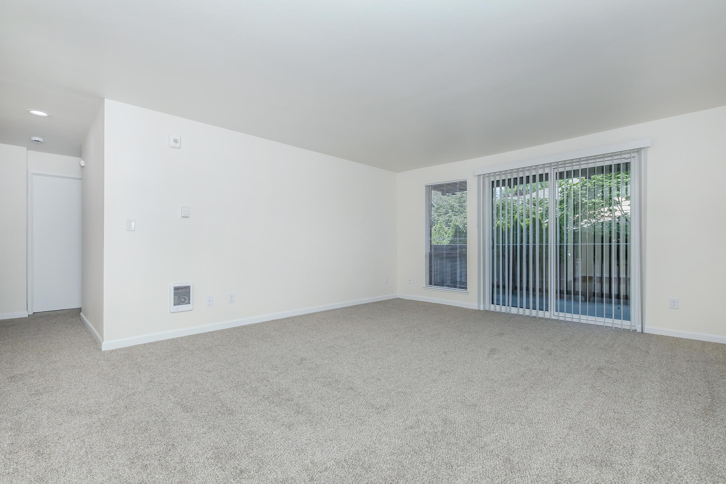 A spacious, empty living room featuring beige carpet and light-colored walls. There is a sliding glass door with vertical blinds that opens to a patio area, and a small vent in the wall. The room is well-lit with natural light coming in from outside.