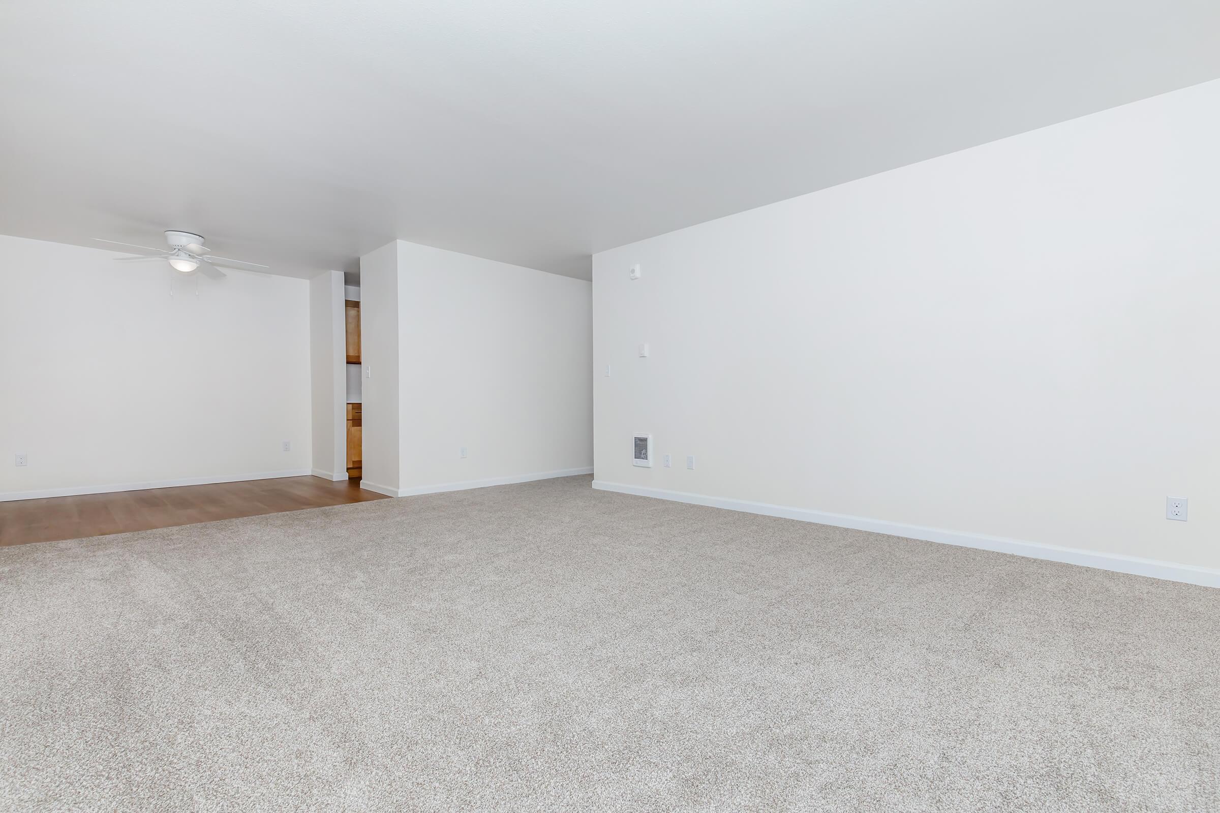 A spacious, empty living room featuring light-colored walls, a ceiling fan, and beige carpet. The room has a seamless transition to another area with hardwood flooring visible in the background. Natural light may flow in through unseen windows, creating a bright and airy atmosphere.