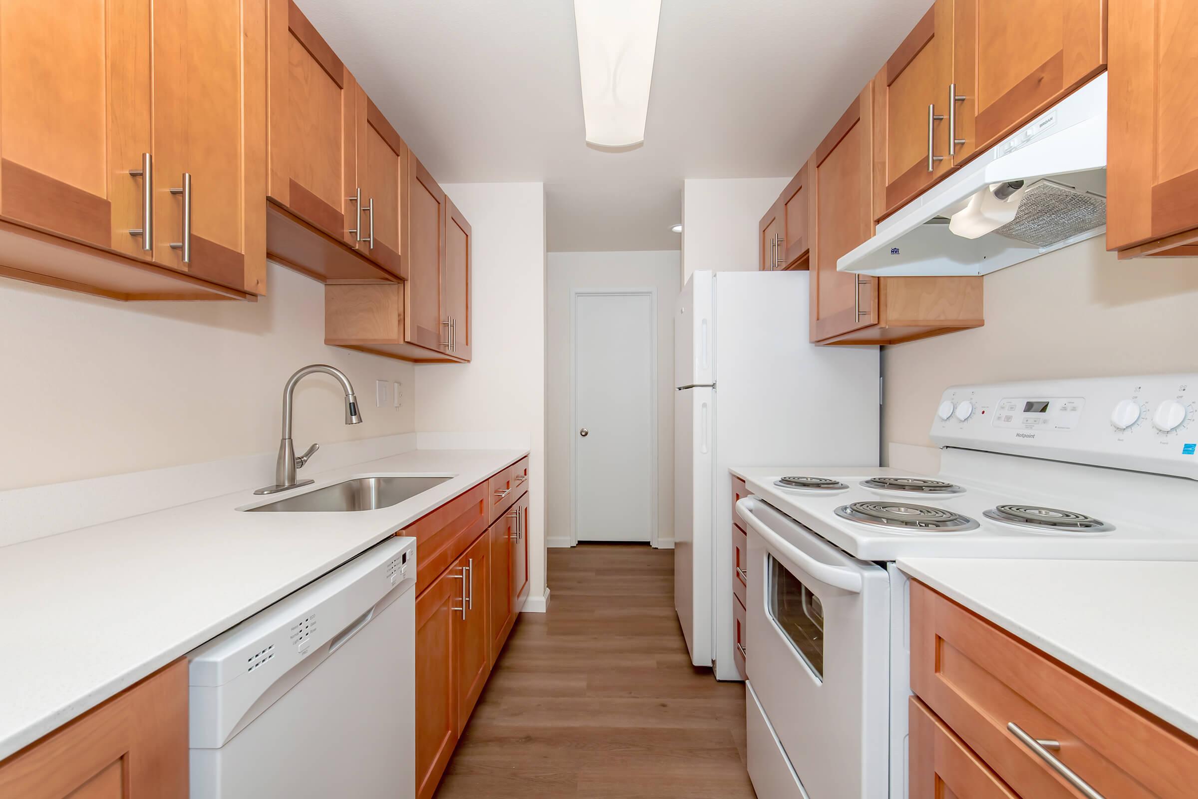A modern kitchen featuring wood cabinets, a stainless steel sink, a dishwasher, a white stove, and a refrigerator. The countertops are light-colored, and the flooring is a warm brown. The layout is clean and functional, with a simple design and ample storage space.