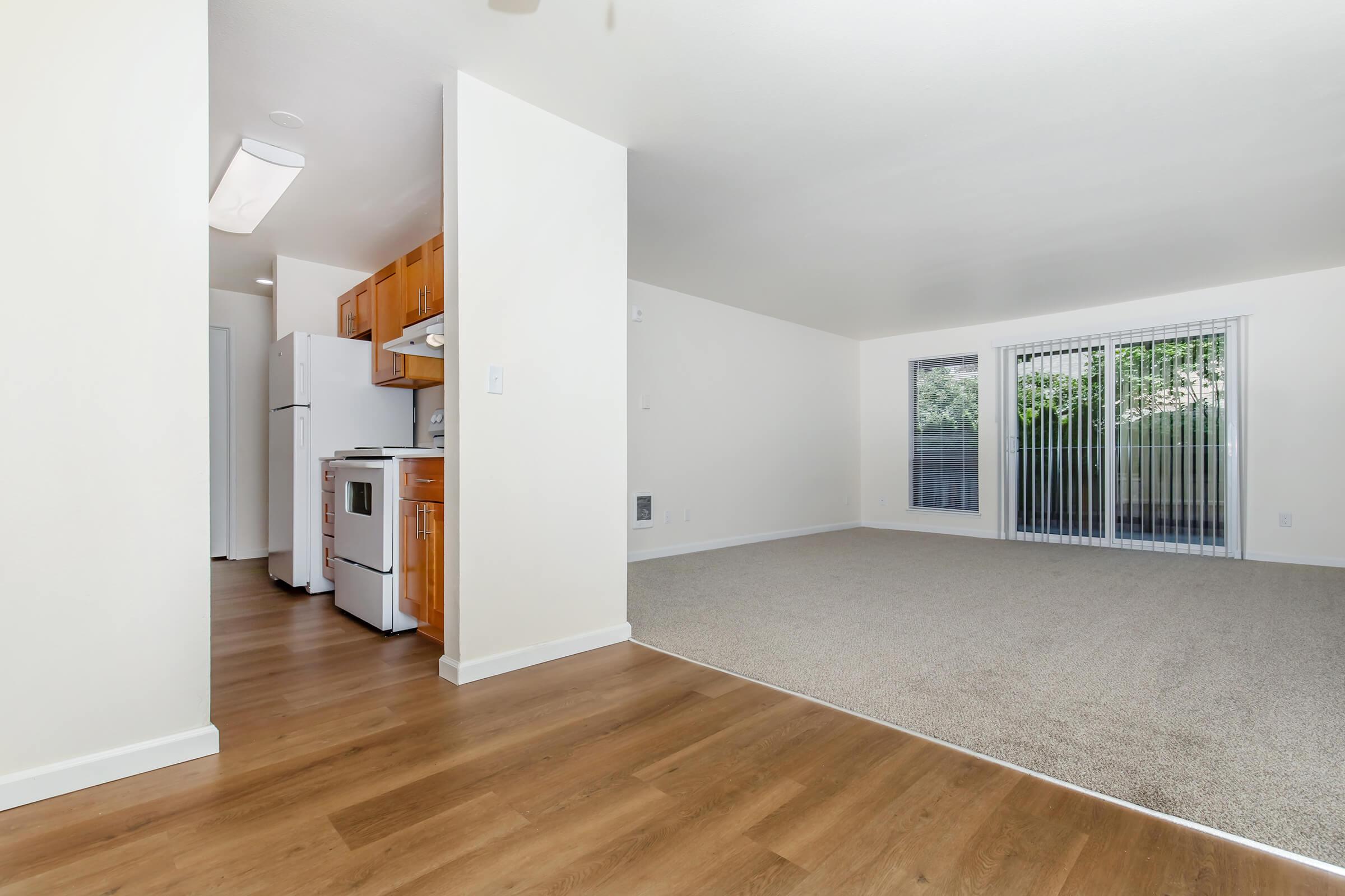A spacious living area with light-colored walls and carpet, featuring a kitchen entry on the left with white appliances. The room has large windows allowing natural light, with a view of greenery outside.