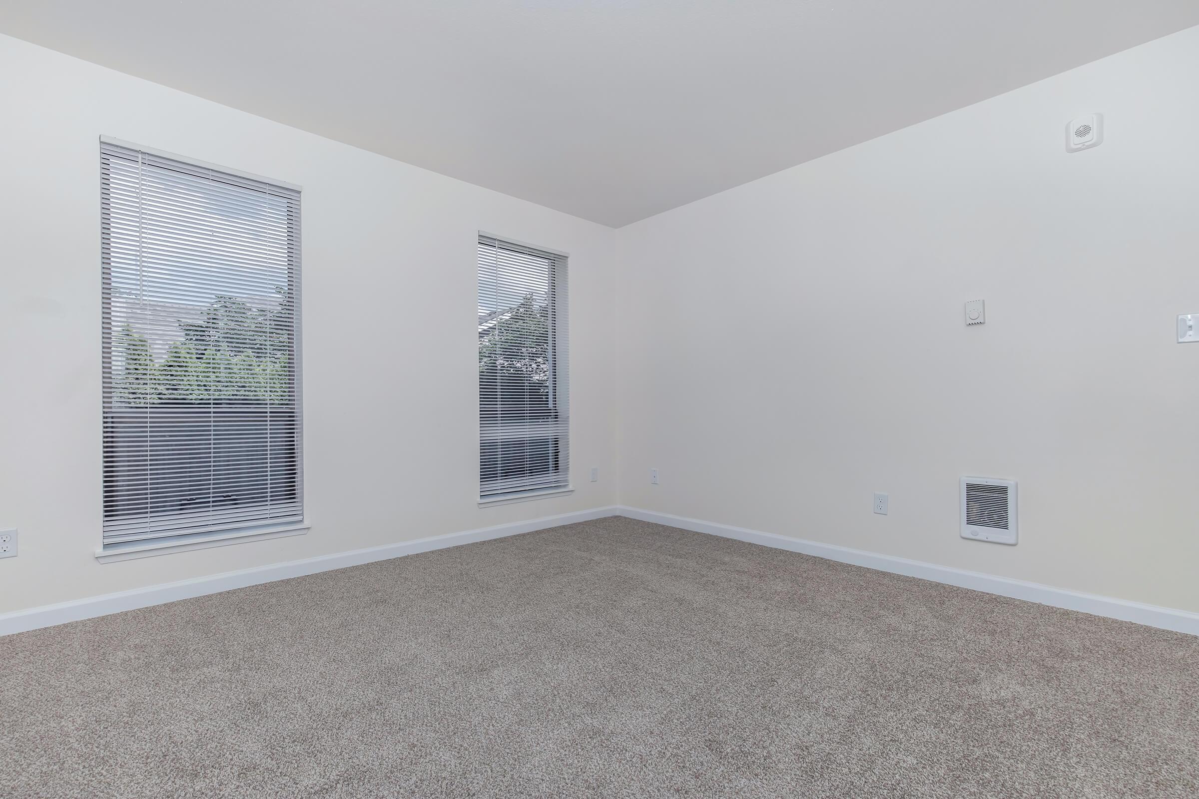 A bright, empty room with two large, tall windows featuring blinds on the left side. The walls are painted white, and the carpet is light beige. A small vent is visible on the right wall, while a single wall switch is located near the corner. The atmosphere is clean and spacious, suitable for furnishing.