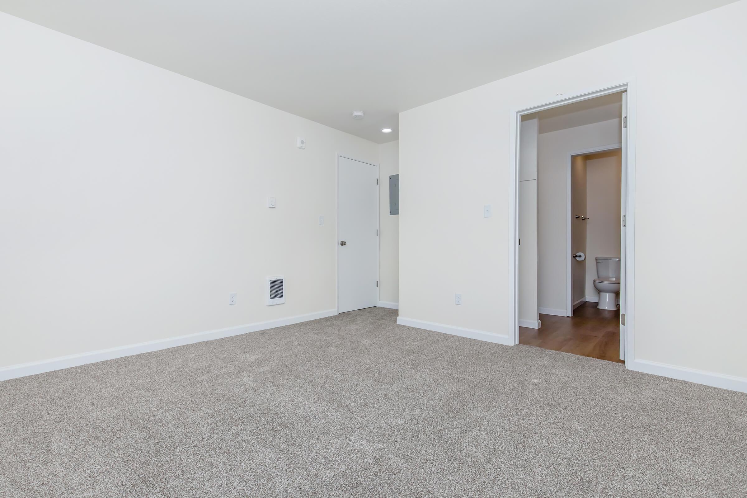 A spacious, empty room with light beige walls and a soft, beige carpet. A closed door on the left leads to the outside, while an open doorway on the right reveals a small bathroom. The room is well-lit with natural light, creating a clean and inviting atmosphere.