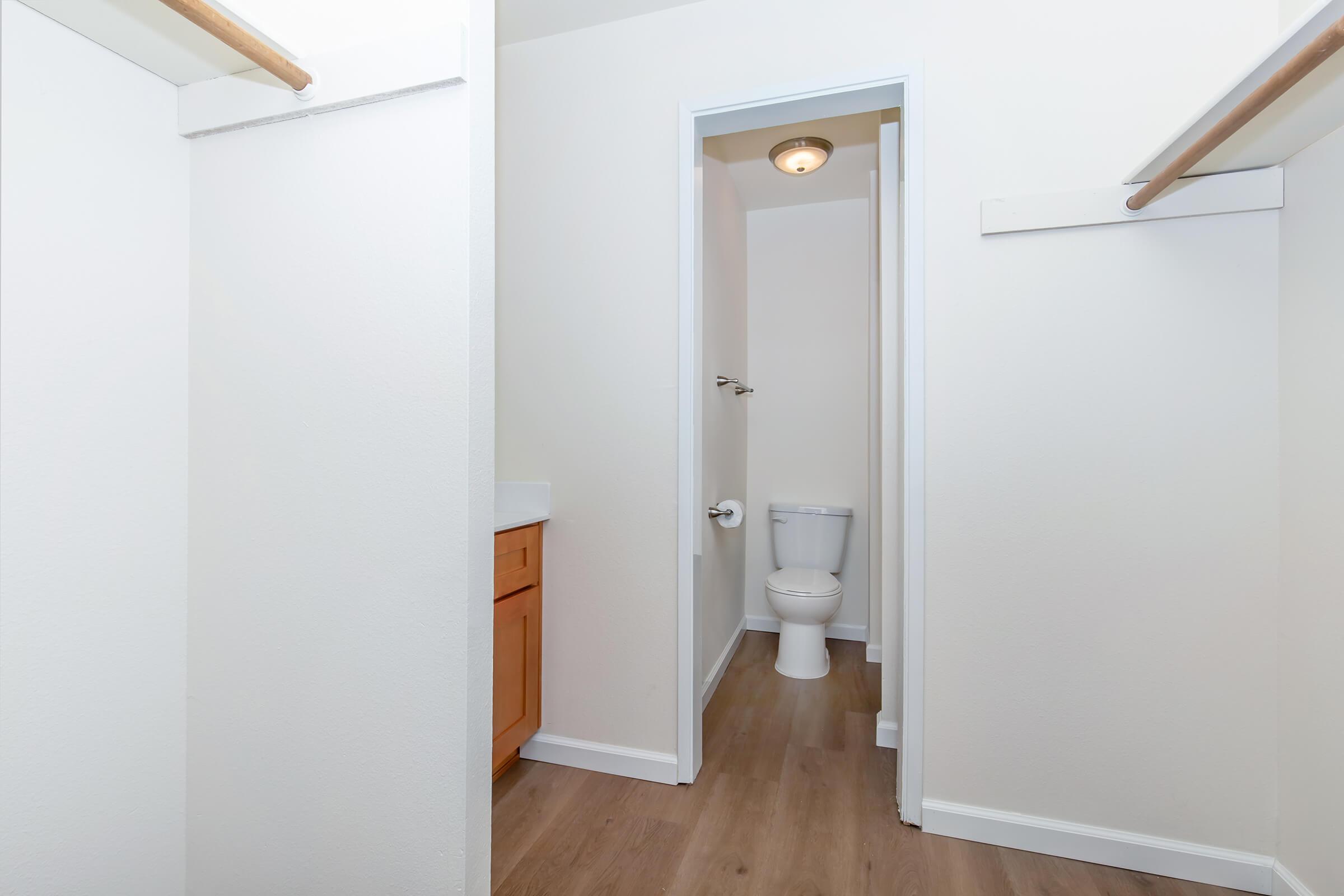 A clean, minimalist bathroom with light-colored walls and wooden flooring. The image features a doorway leading to a small restroom that includes a toilet. A wooden cabinet is visible near the entrance, with a towel rack on the wall and ample space, creating a bright and open atmosphere.