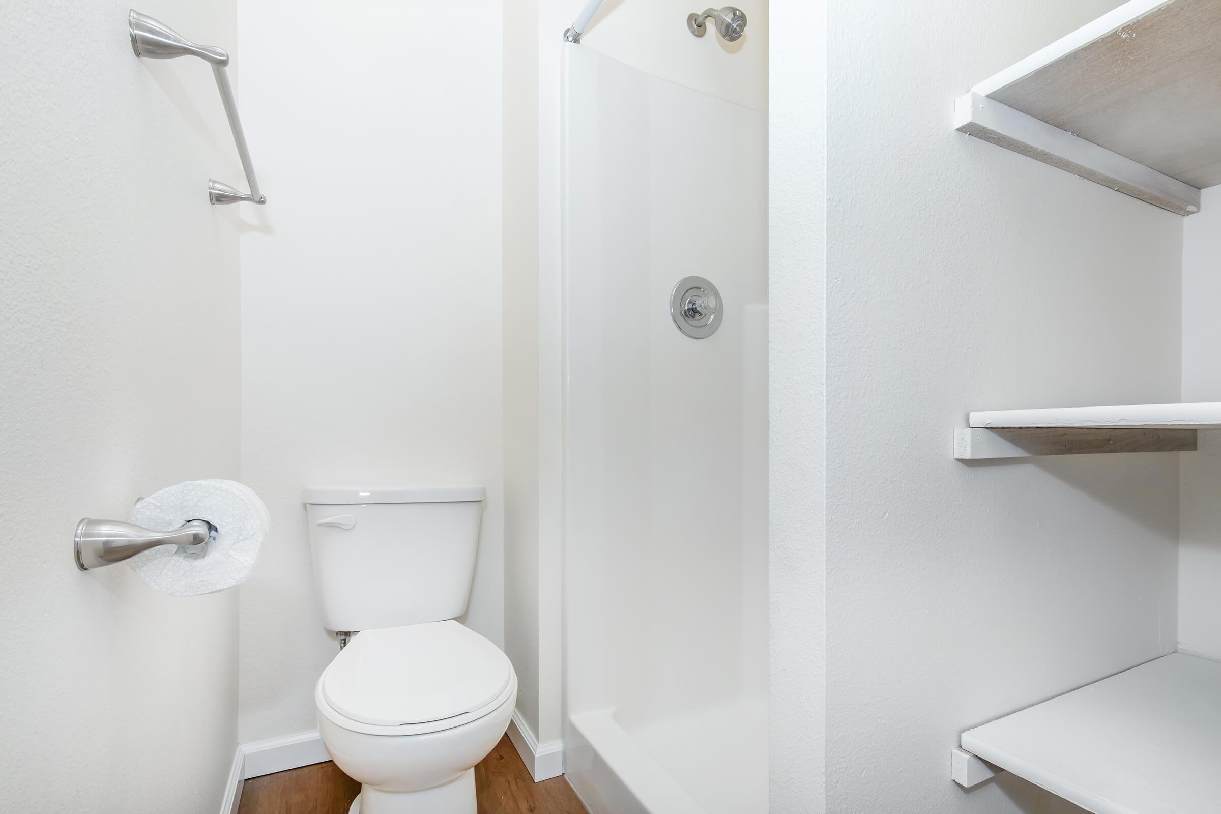 A small bathroom featuring a white toilet, a simple glass shower enclosure, a toilet paper holder, and a shelf on the wall. The walls are painted in a light color, and the floor is wooden, creating a clean and modern look.