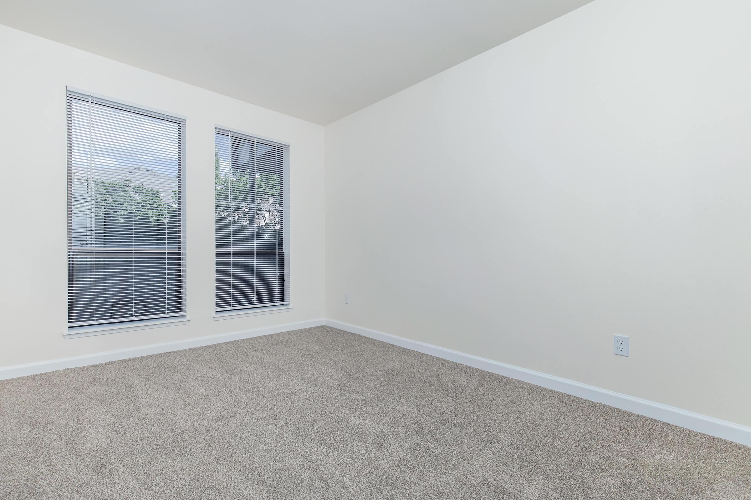 Empty room with light beige carpet and white walls. Two large windows with closed blinds allow natural light to enter, illuminating the space. The room is devoid of furniture, creating a clean and minimalistic aesthetic.