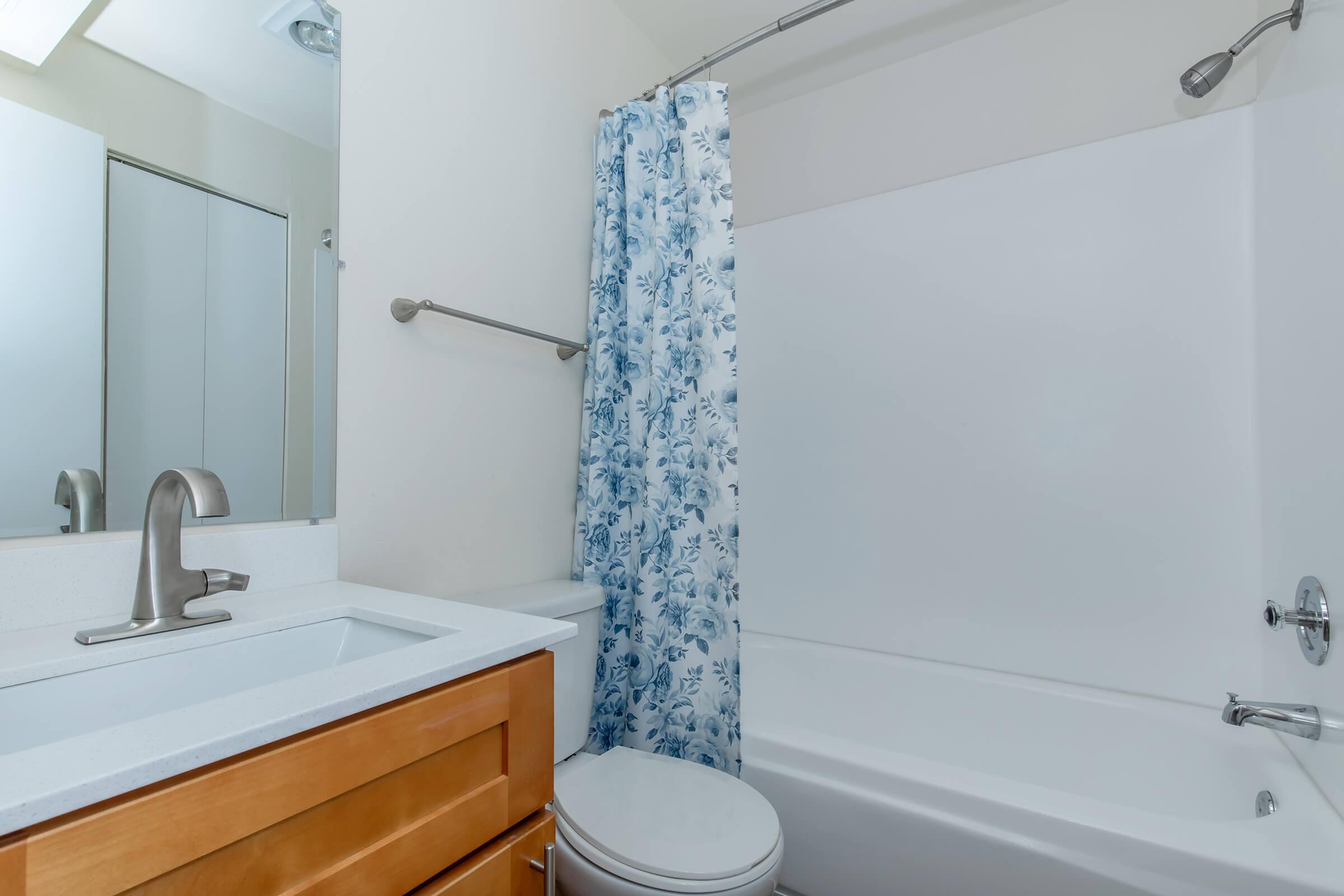 Bathroom interior featuring a bathtub with a blue floral shower curtain, a white sink with a chrome faucet, and a toilet. Soft, neutral colors dominate the space, with a large mirror reflecting light. Bright and clean ambiance, showcasing modern fixtures and simple decor.