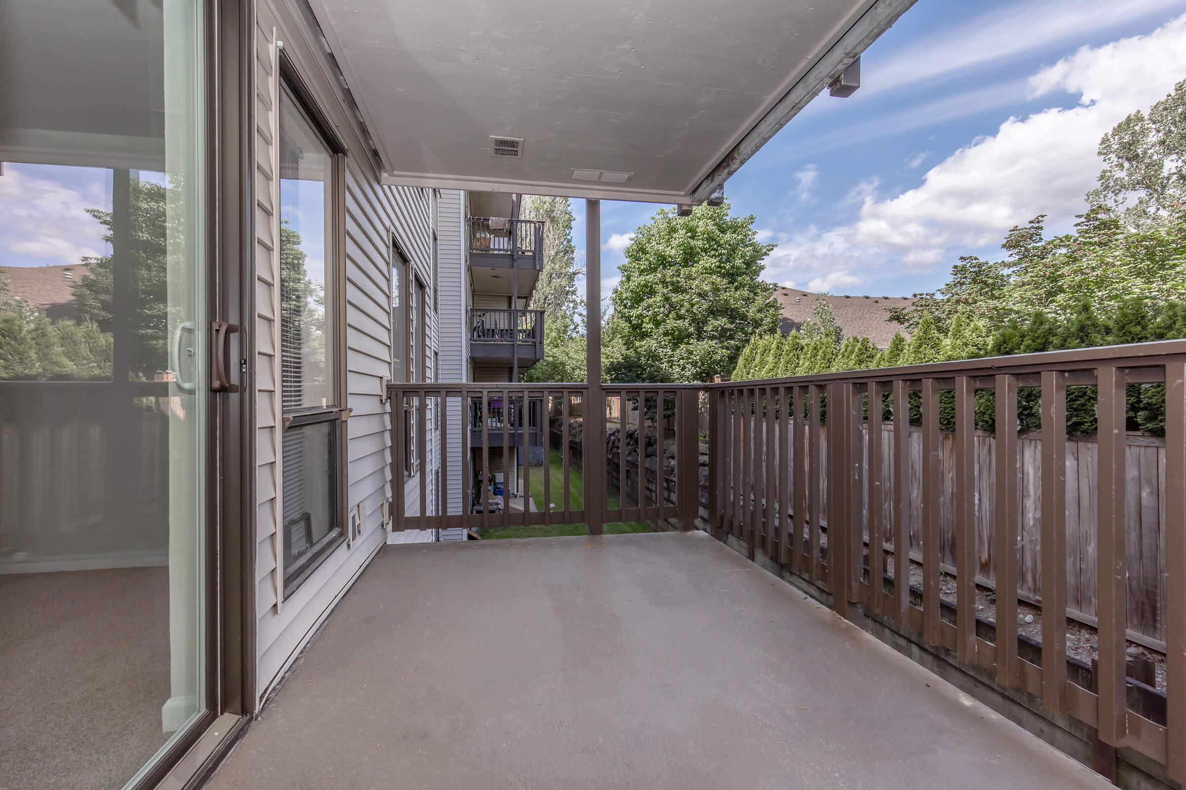 A spacious balcony with a wooden railing, featuring a view of trees and nearby buildings under a partly cloudy sky. The floor is concrete, and there are sliding glass doors leading to an indoor space.